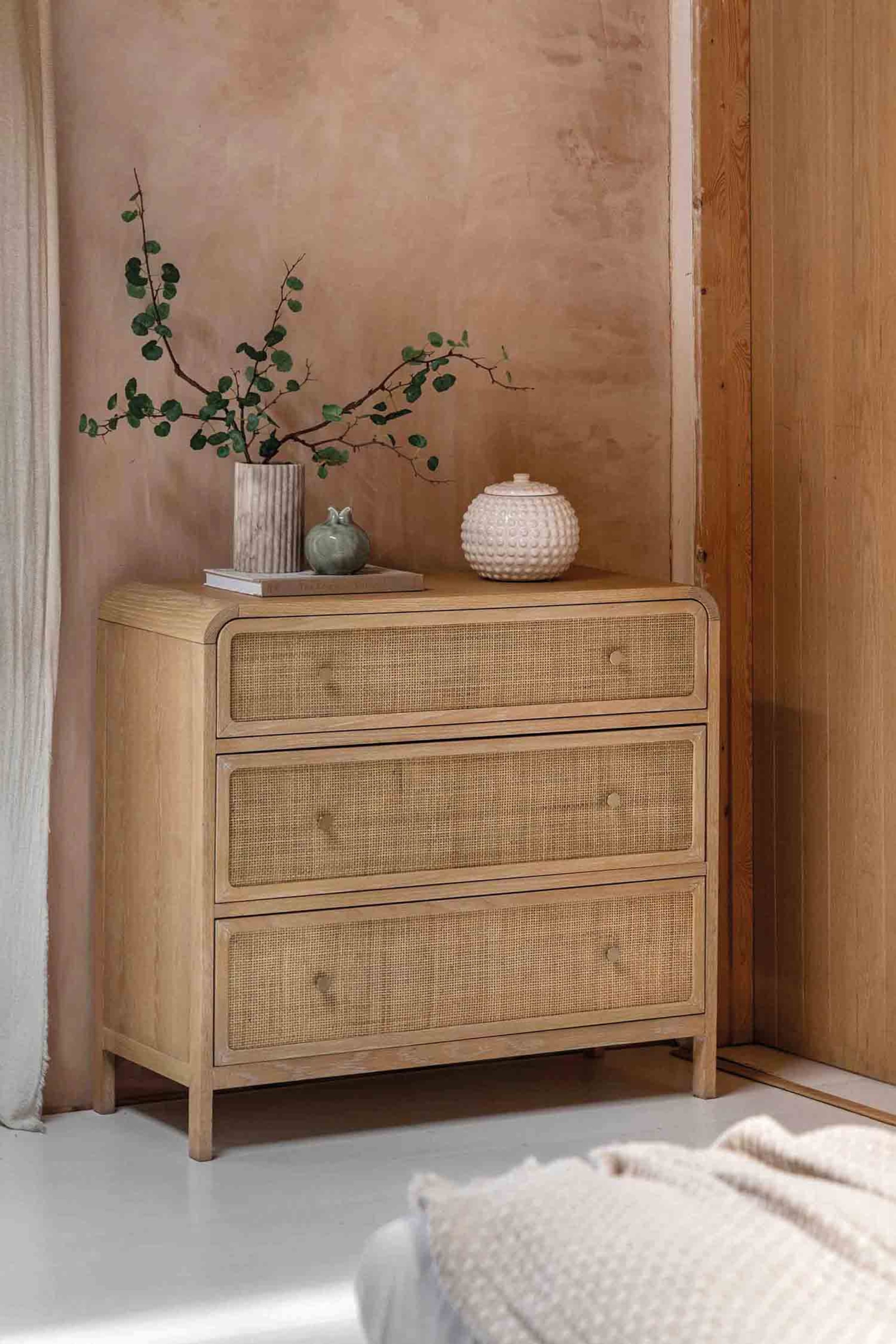 View larger image: A light-toned wooden chest of drawers is showcasing its front and left side, featuring three drawers with natural cane webbing inserts. Each drawer is adorned with a small, round knob, and the chest stands on short, tapered legs. Its top edges curve gently.