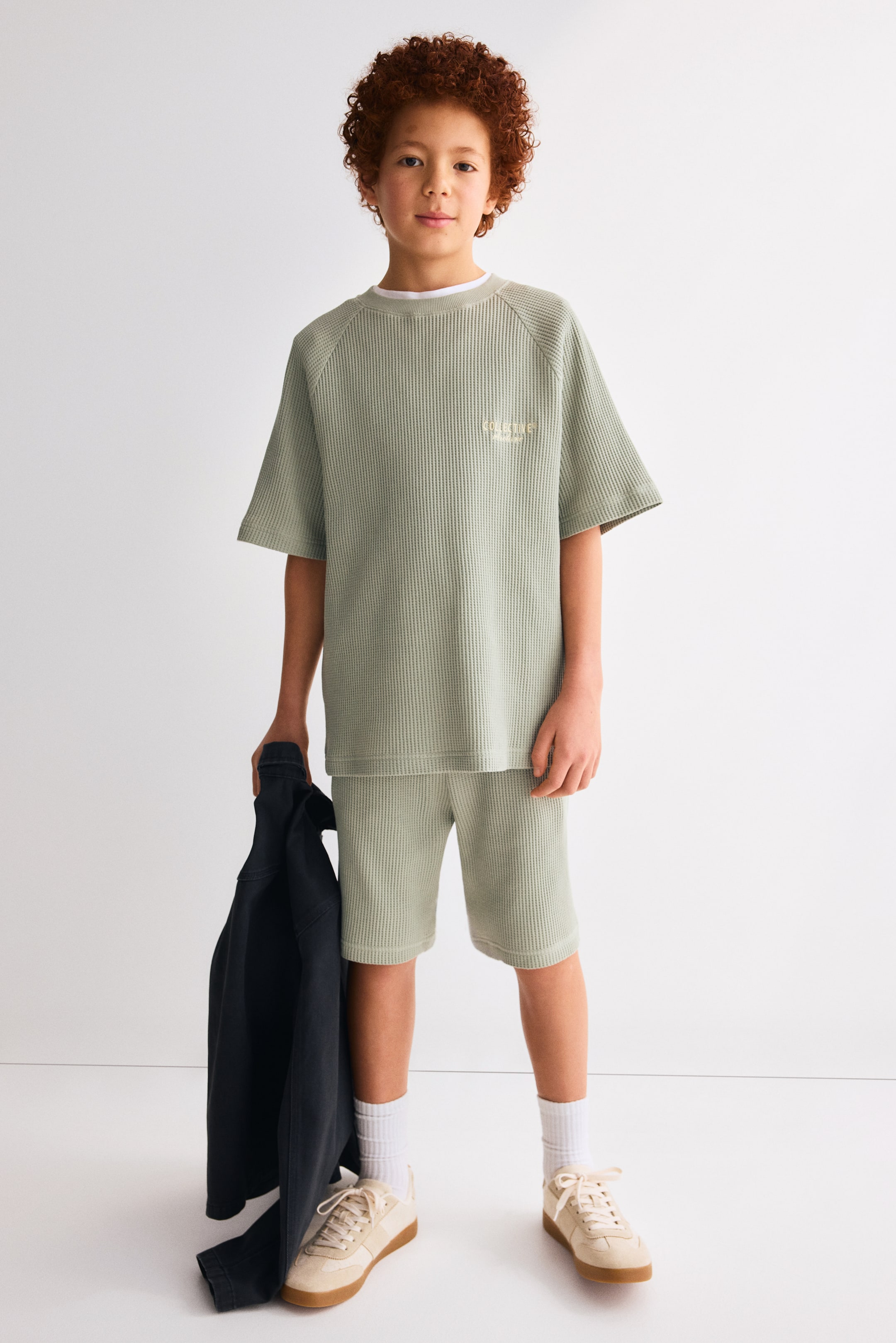 Visualizza immagine più grande: Un completo composto da una t-shirt e pantaloncini in tessuto waffle di colore verde salvia è presentato frontalmente. La t-shirt a maniche corte presenta maniche raglan, un girocollo con una maglietta bianca visibile sotto, e un logo ricamato in marrone chiaro sul petto sinistro, mentre i pantaloncini coordinati arrivano sopra il ginocchio.