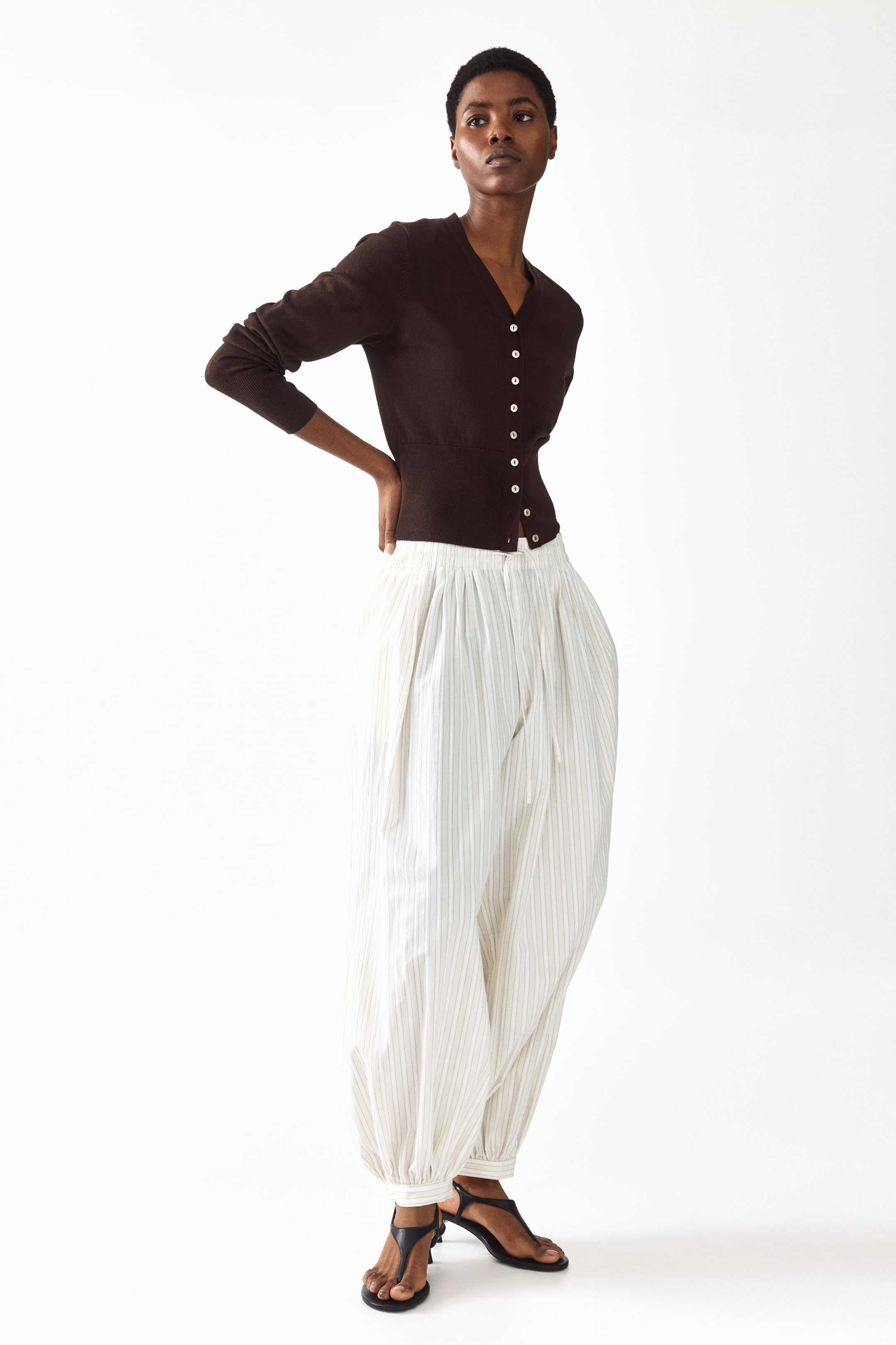 Zobraziť väčší obrázok: Modelka má na sebe krátky, priliehavý hnedý jemne pletený kardigán s výstrihom do V, bielymi gombíkmi vpredu a dlhými rukávmi. Má široké, rebrované lemy na manžetách a spodnom okraji. Modelka pózuje s rukami na bokoch, pričom ho nosí na svetlých nohaviciach.