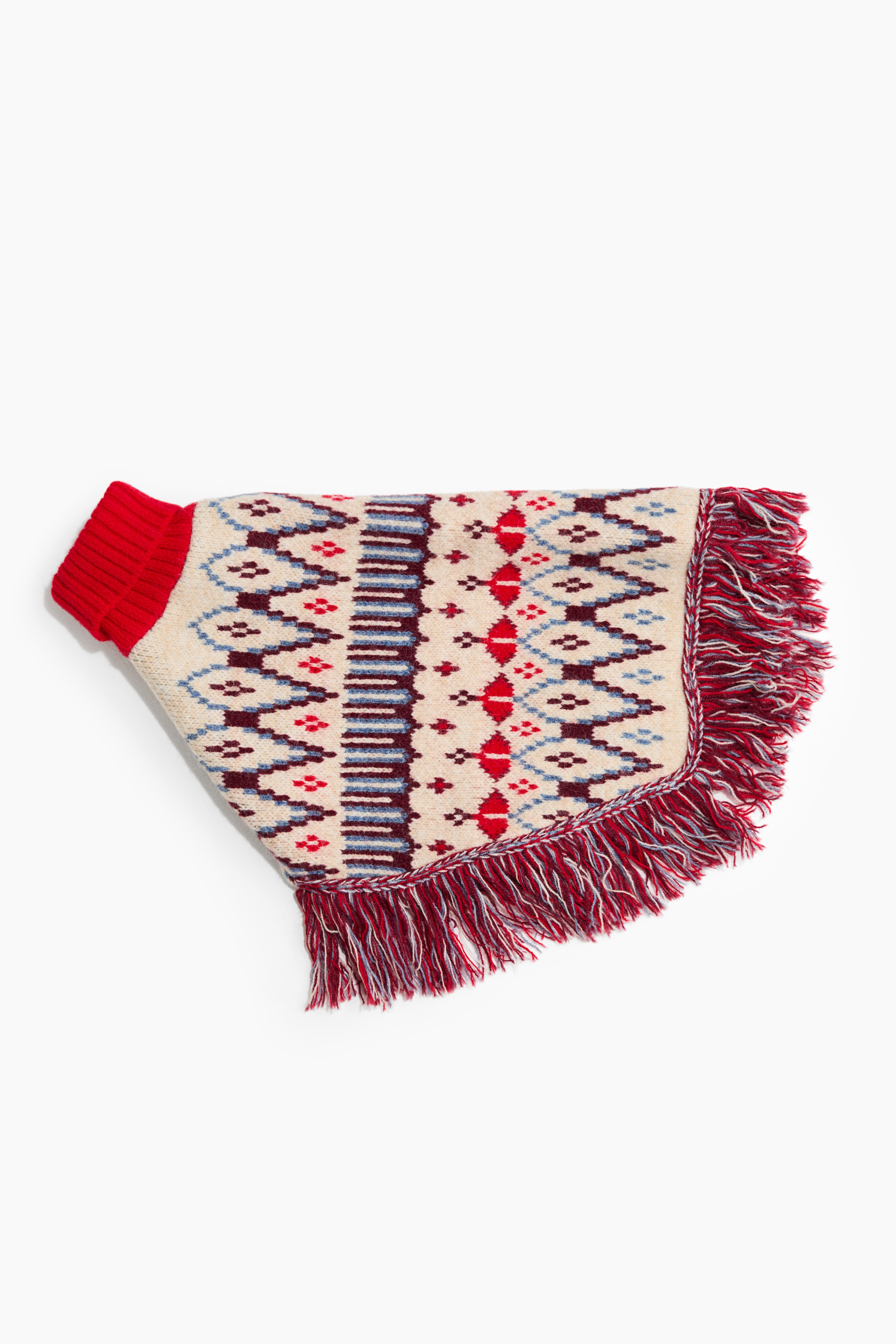 A dog cape in cream-coloured jacquard knit with red and blue geometric patterns is draped over a brown dog facing left, showcasing its fit and design. The cape features a thick, red rib-knit roll neck and a multicoloured fringed trim along the bottom hem.