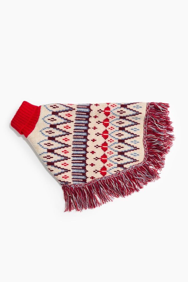 Ein gestrickter Hundeponcho liegt flach und zeigt einen roten, gerippten Rollkragenkragen sowie einen cremefarbenen KÃ¶rper mit einem geometrischen Strickmuster in Rot, Blau und Burgunderrot. Der geschwungene untere Saum ist mit einer dichten, mehrfarbigen Fransenverzierung versehen. - 1