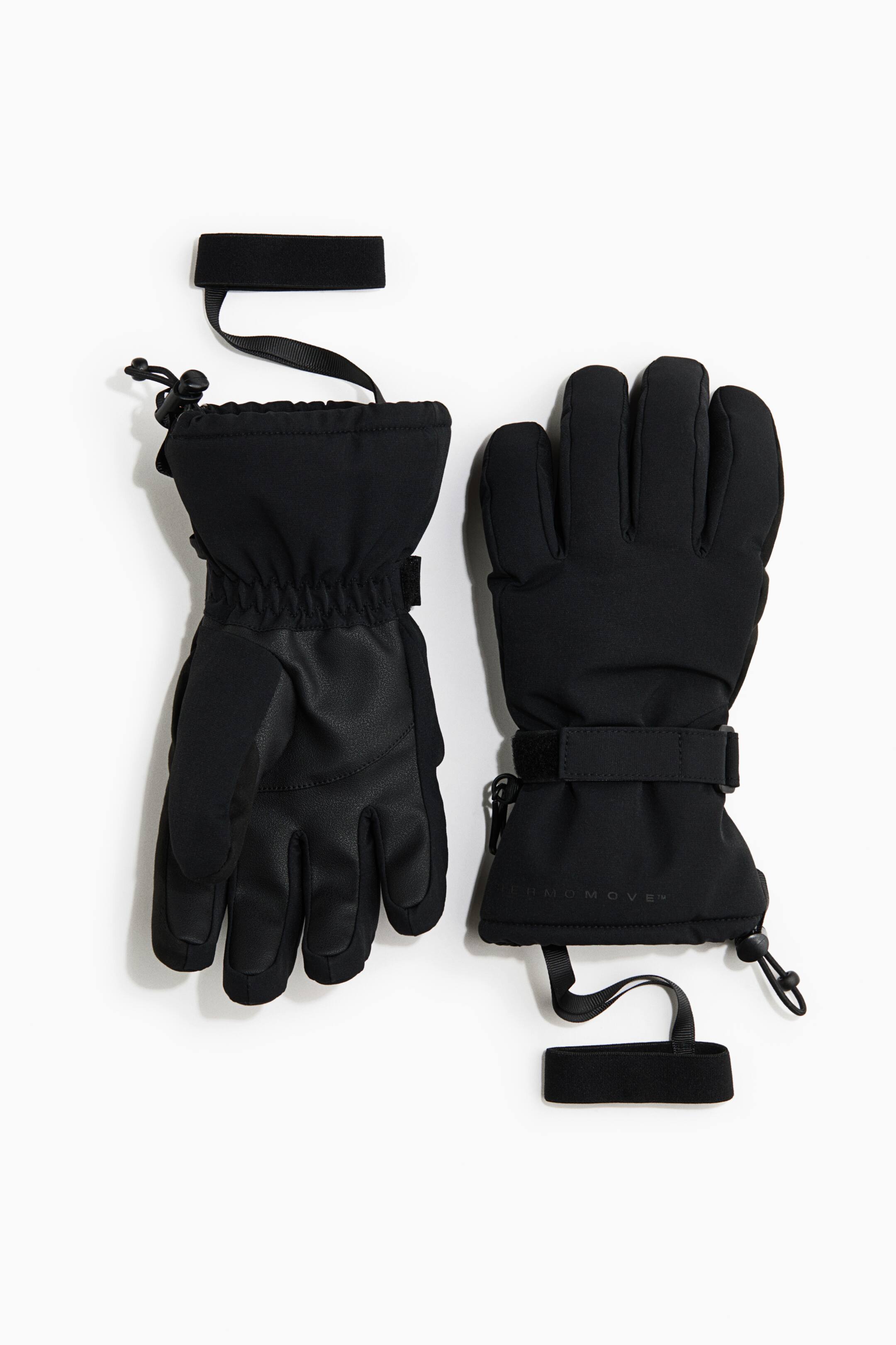 Ampliar la imagen: Un par de guantes negros de invierno está colocado en posición horizontal, con un guante mostrando la palma texturizada de agarre y el otro mostrando el dorso liso de la mano. Ambos guantes cuentan con correas ajustables en la muñeca, cordones con ajuste en los puños tipo guantelete y correas elásticas de seguridad integradas.