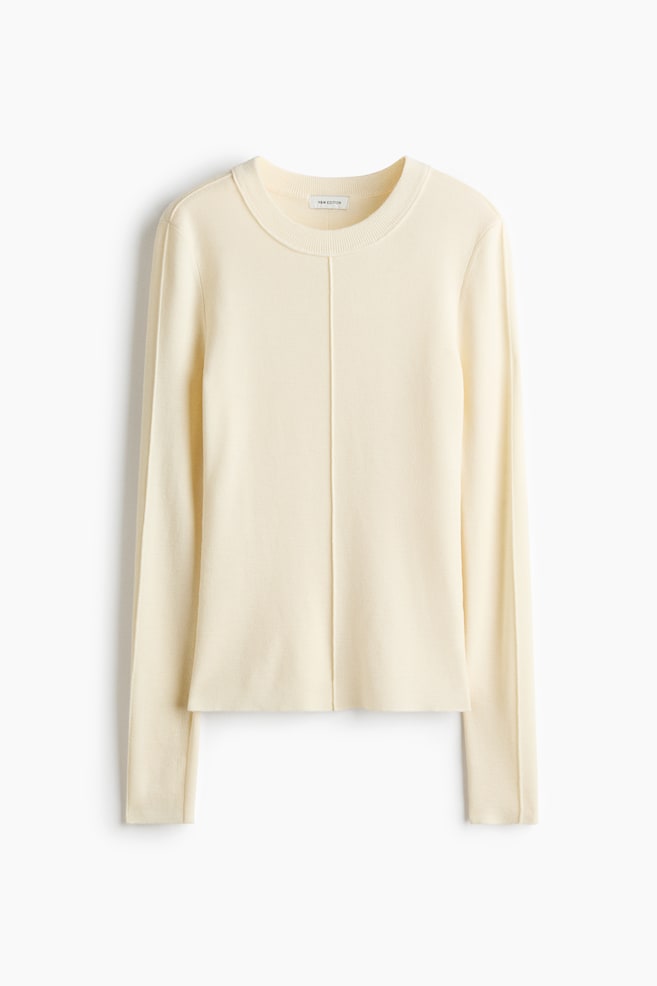 A cream-coloured merino wool knitted top is shown from the front, featuring a round, rib-trimmed neckline and long sleeves. Distinctive linked seams run vertically down the centre front of the top and along the outer length of each sleeve. - 1