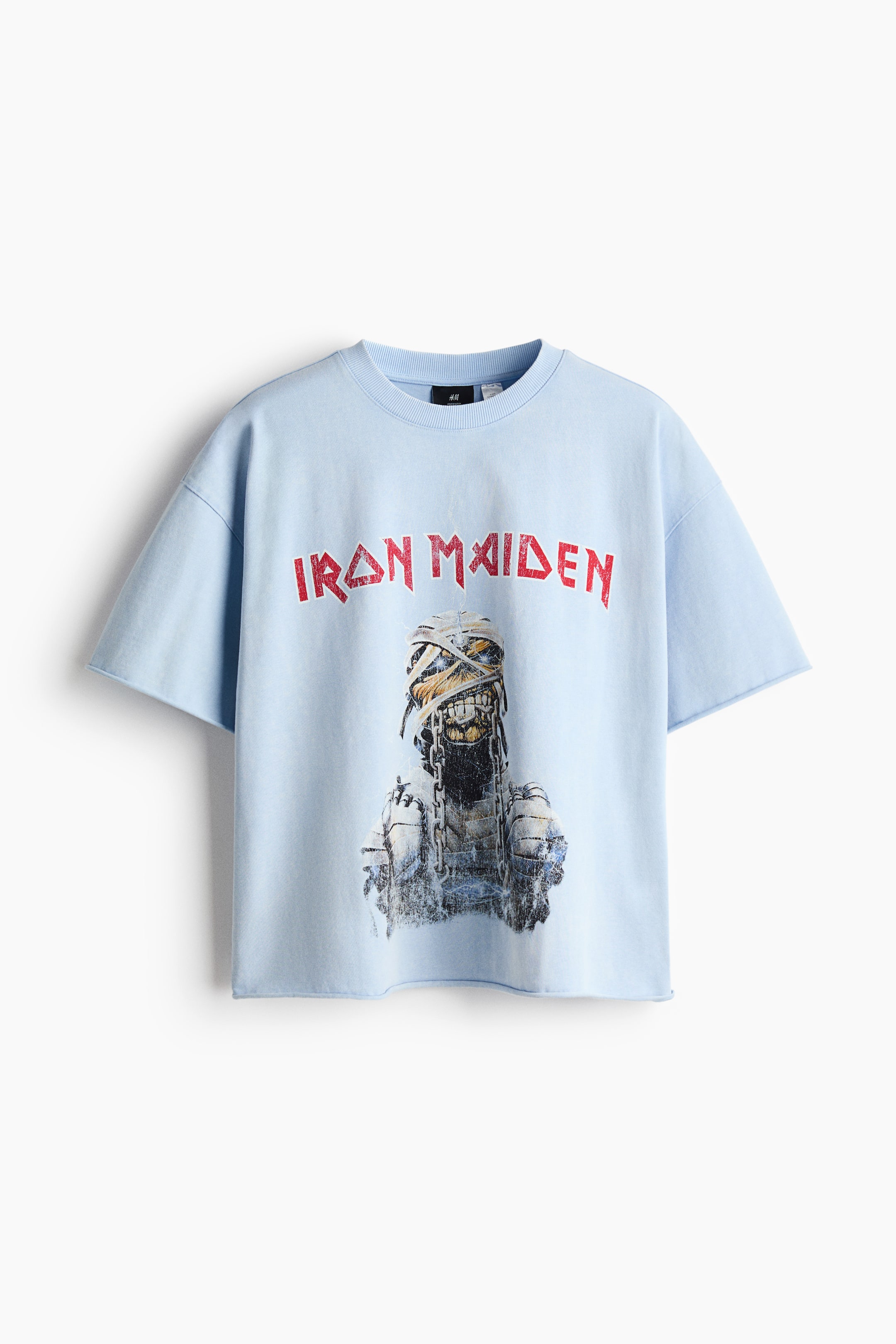Ver imagen más grande: Una camiseta de manga corta azul claro está extendida, vista de frente, mostrando un texto impreso en rojo "IRON MAIDEN" sobre un gráfico de una figura tipo momia con cadenas. Presenta un cuello redondo acanalado, mangas cortas con corte sin rematar y un dobladillo inferior con corte sin rematar.