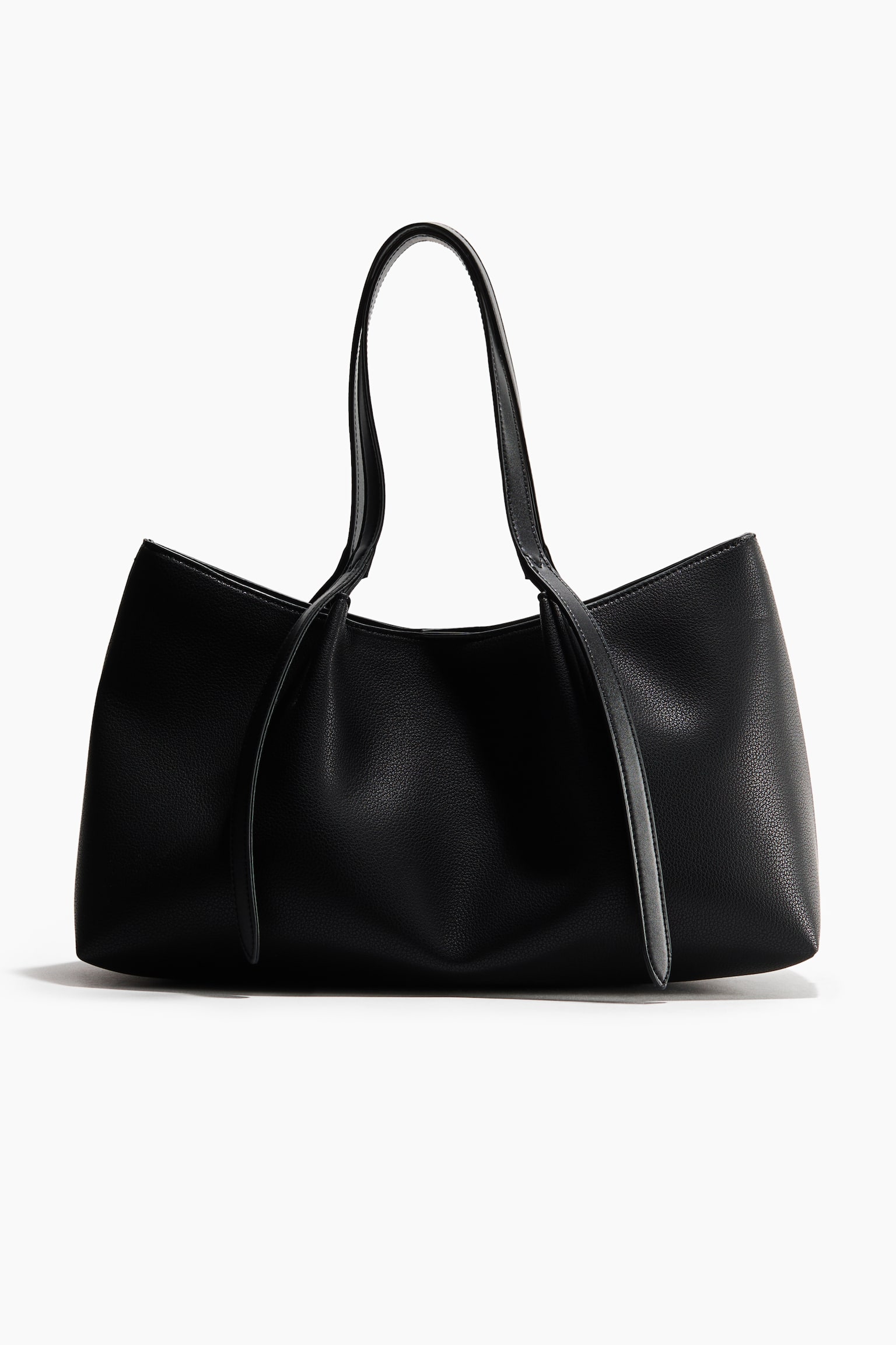Un bolso tote de cuero negro con acabado granulado aparece de frente, mostrando su estructura suave y holgada y su acabado texturizado. Presenta dos asas superiores finas con largos extremos de correa planos que caen por la parte frontal del bolso.