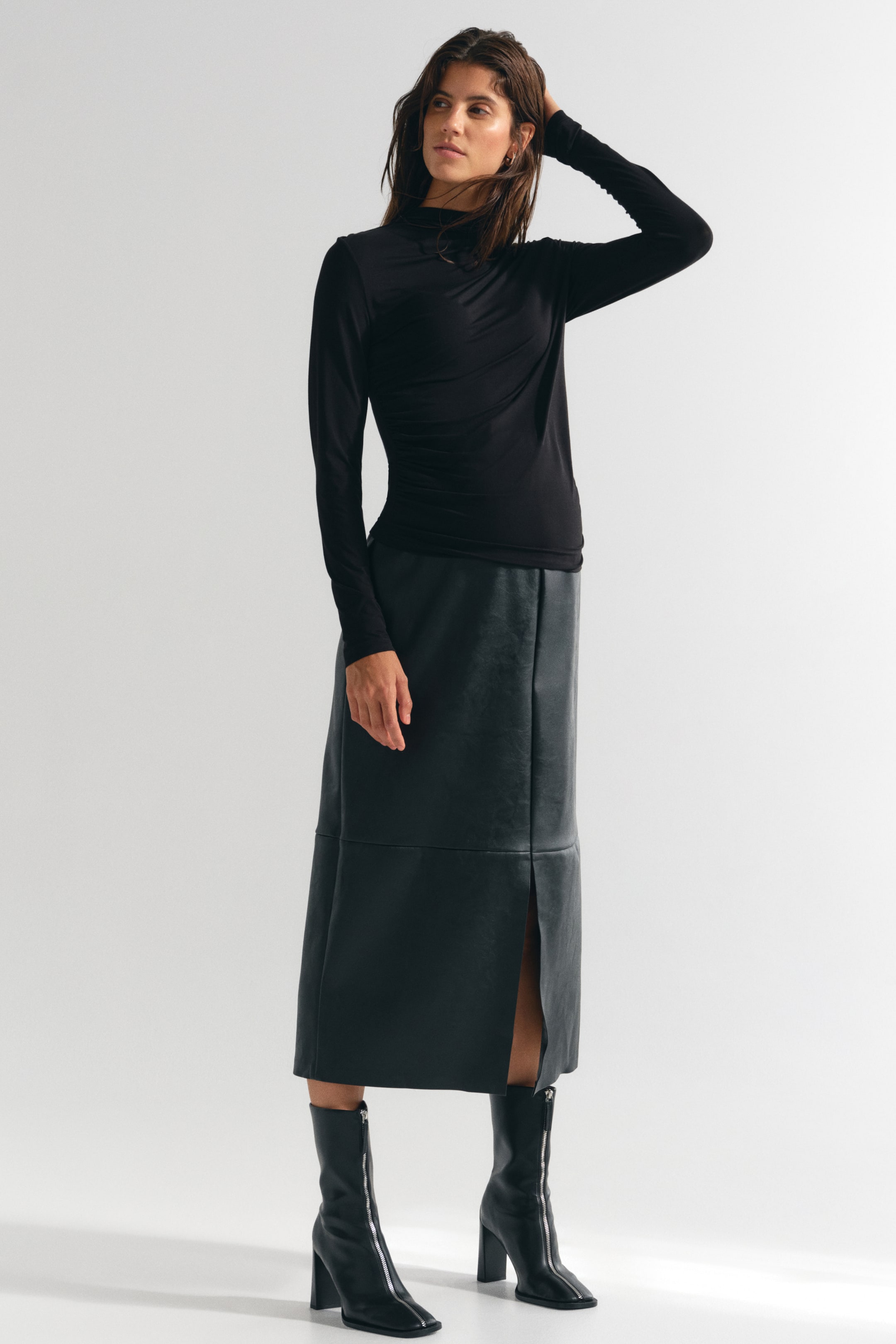 View larger image: A black calf-length skirt in a coated fabric is being worn by the model, featuring prominent horizontal and vertical seams and a high front slit. The skirt is styled with a black long-sleeved top and black high-heeled boots.