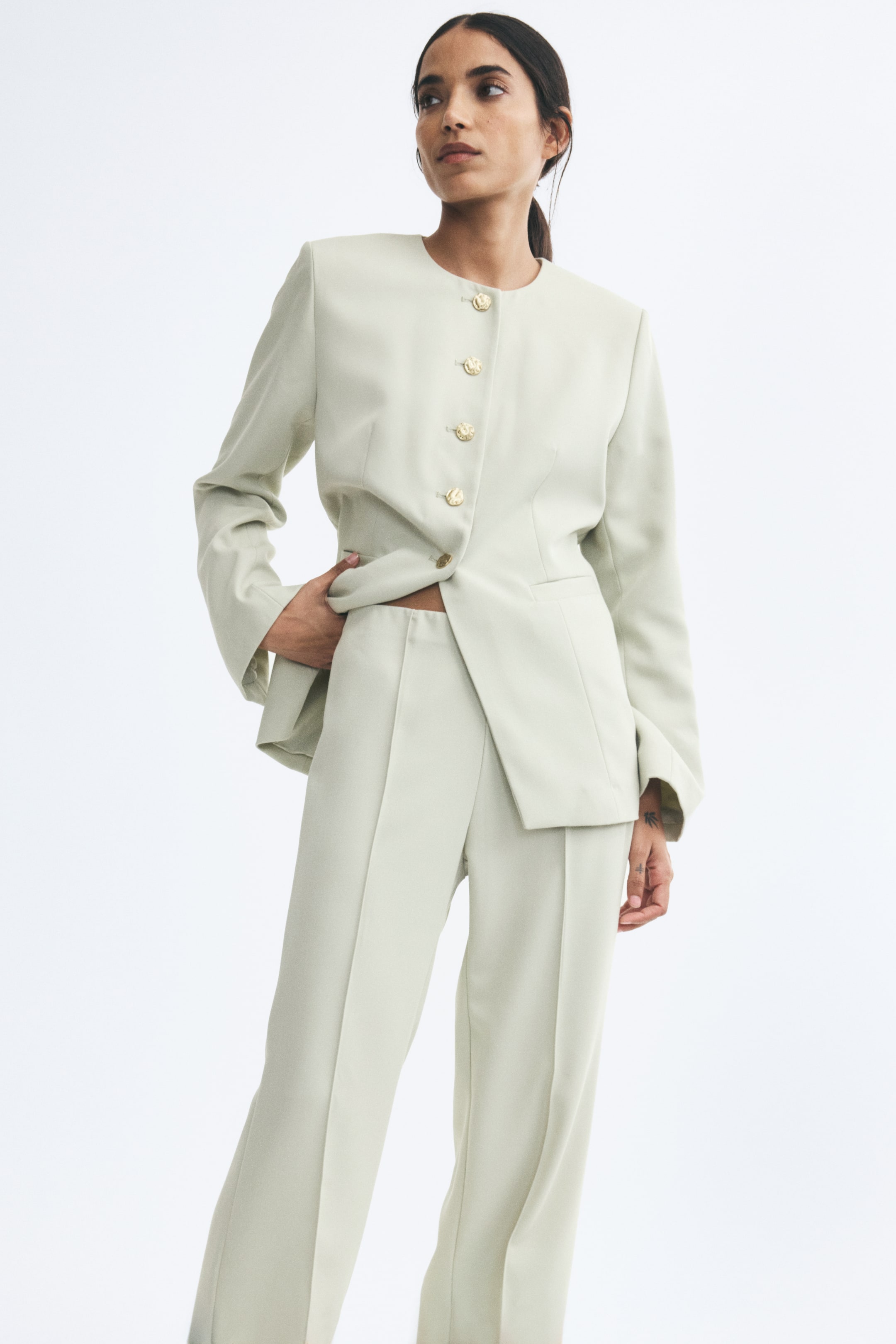 View larger image: The model is wearing light green fitted twill trousers featuring a high waist and crisp front creases. The trousers are styled with a matching collarless blazer, with the model resting one hand near the waistband and slightly lifting the jacket hem.