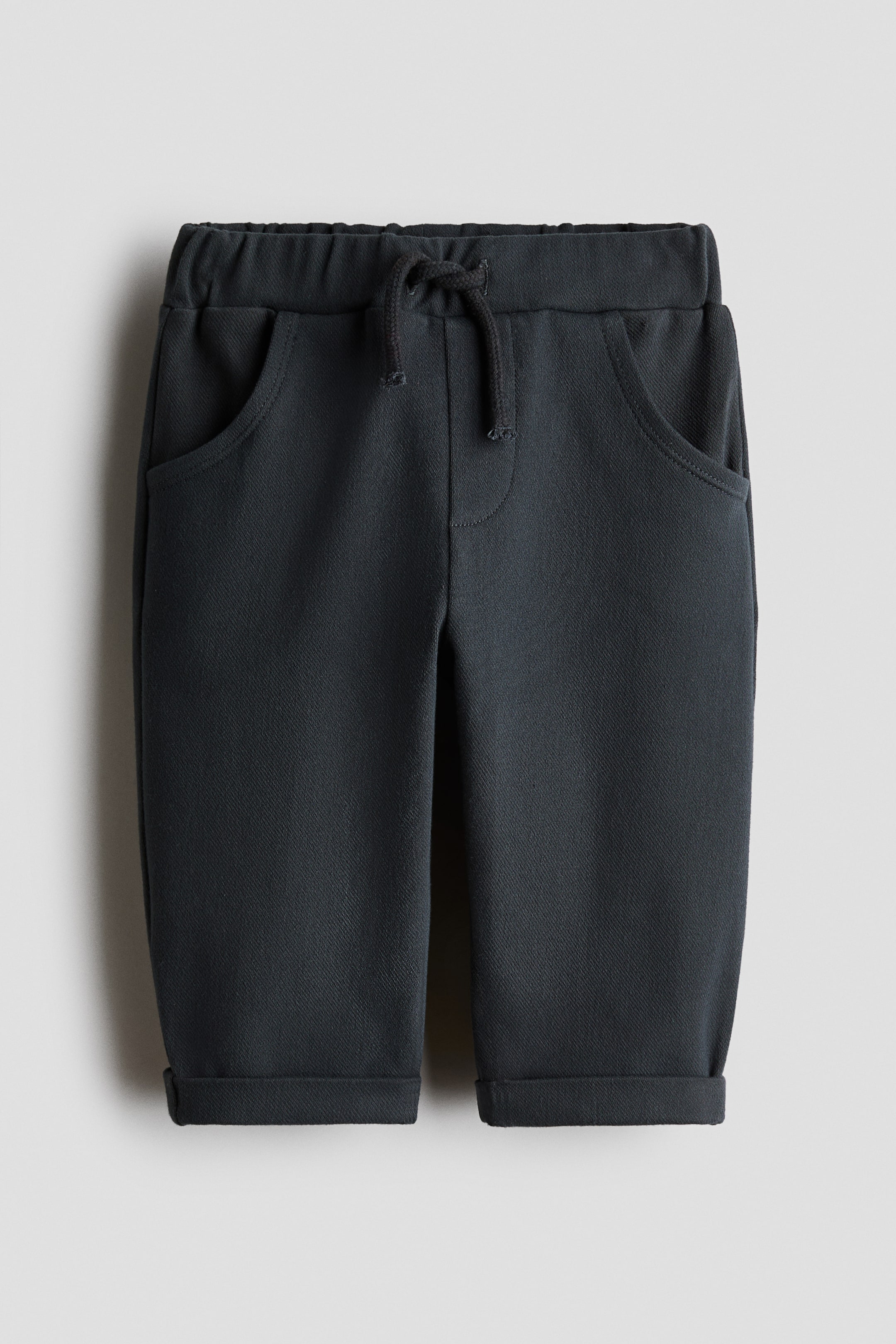 View larger image: Trousers in dark grey denim-look cotton jersey are showcased from the front, featuring an elasticated waist with a decorative drawstring, a faux fly, and curved front pockets. Sewn-in turn-ups complete the hems.