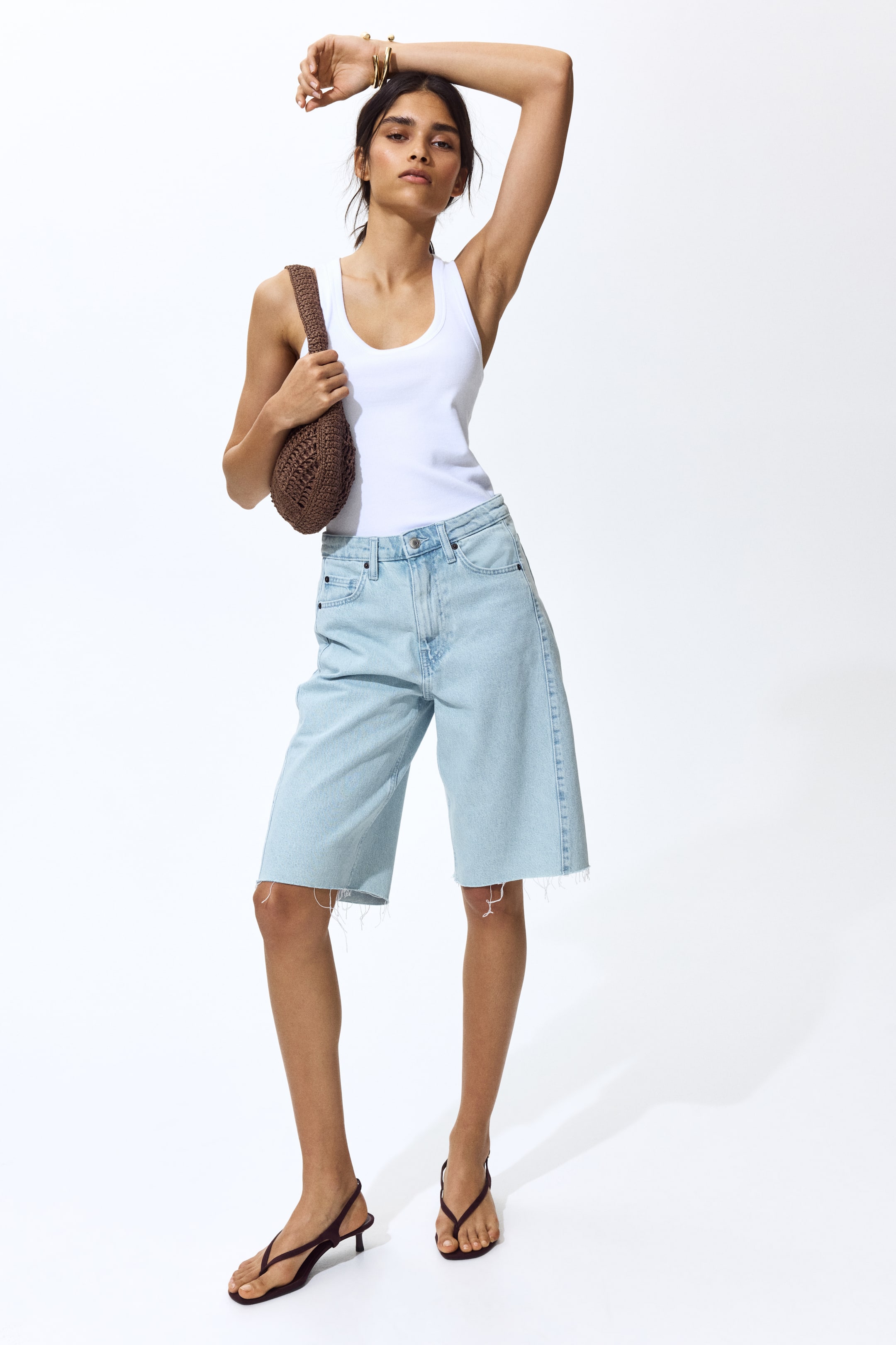 Grösseres Bild ansehen: Knielange, 5-Pocket-Shorts aus festem hellblauen Baumwoll-Denim, mit hoher Taille, Reissverschluss und Knopf sowie rohen Säumen, werden vom Model getragen. Das Model trägt zudem ein weisses Tanktop, das in die Shorts gesteckt ist.