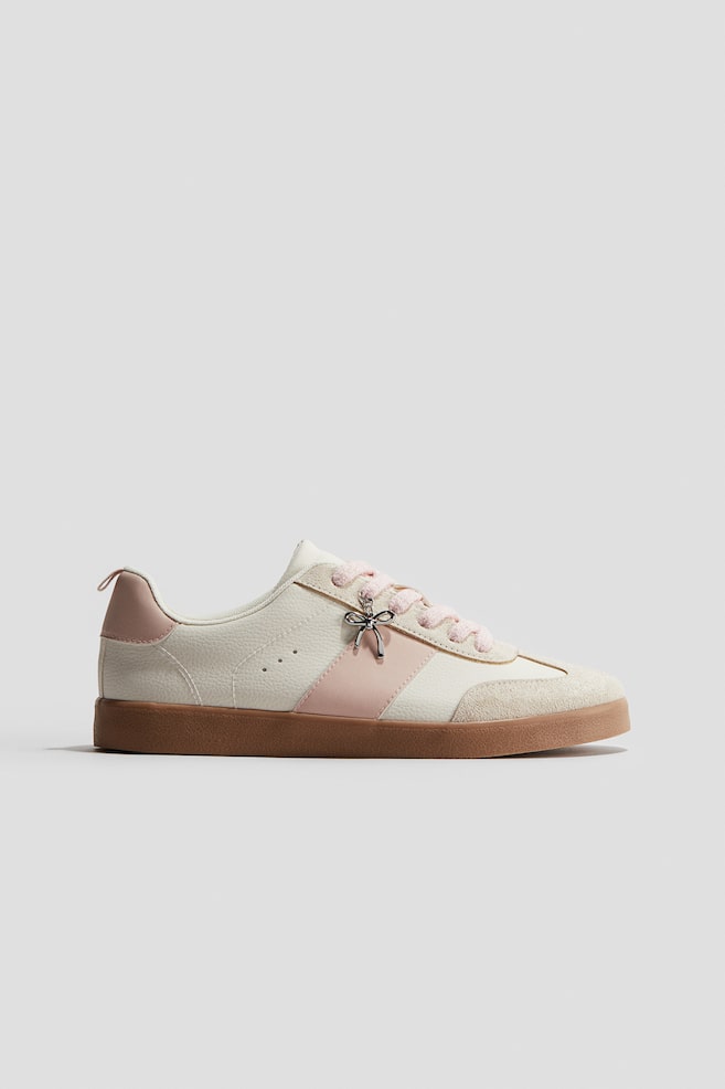 An off-white and pink sneaker is presented in a side view. It is featuring textured off-white panels, an off-white suede toe cap, and light pink accents on the side stripe and heel counter. Light pink laces are threaded through eyelets, adorned with a silver-toned bow charm, and the sneaker is resting on a brown gum sole. - 1
