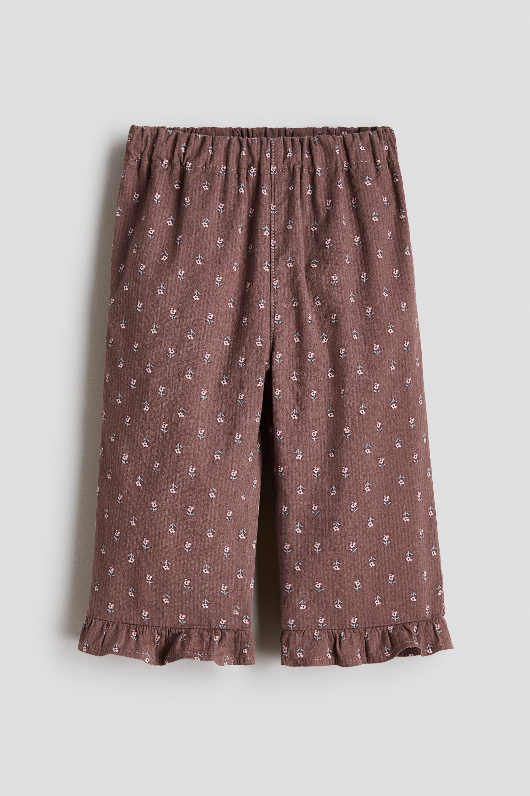 View larger image: These pants are shown from the front, crafted from a muted reddish-brown fabric with a subtle vertical texture and a small floral print in light pink and blue throughout. They feature an elasticated, gathered waist, straight legs, and frill-trimmed hems.