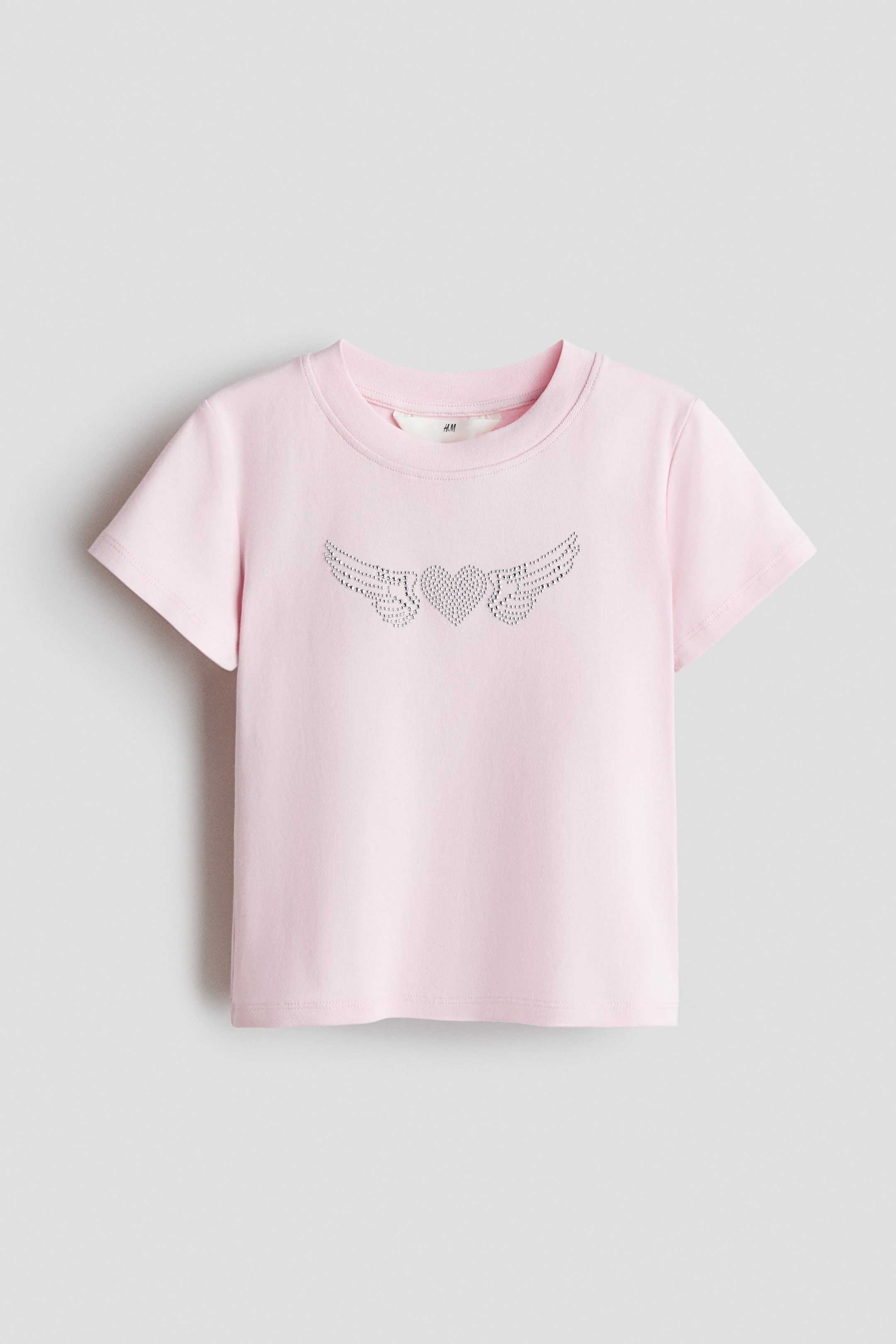 查看大圖: 粉紅色的 T 恤採用柔軟的棉質針織面料製成，領口周圍有羅紋飾邊，正面展示了裝飾心形和翅膀圖案。