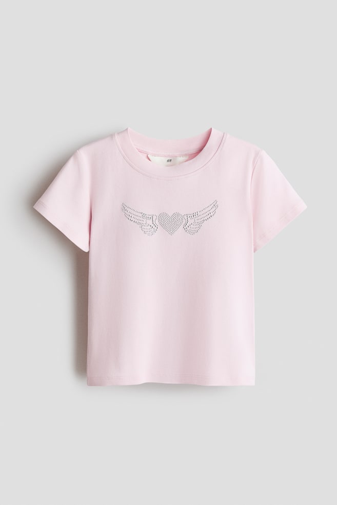 Un t-shirt rose en jersey de coton doux, avec une bordure côtelée autour de l'encolure, arborant un motif de cœur orné et d'ailes sur le devant. - 1