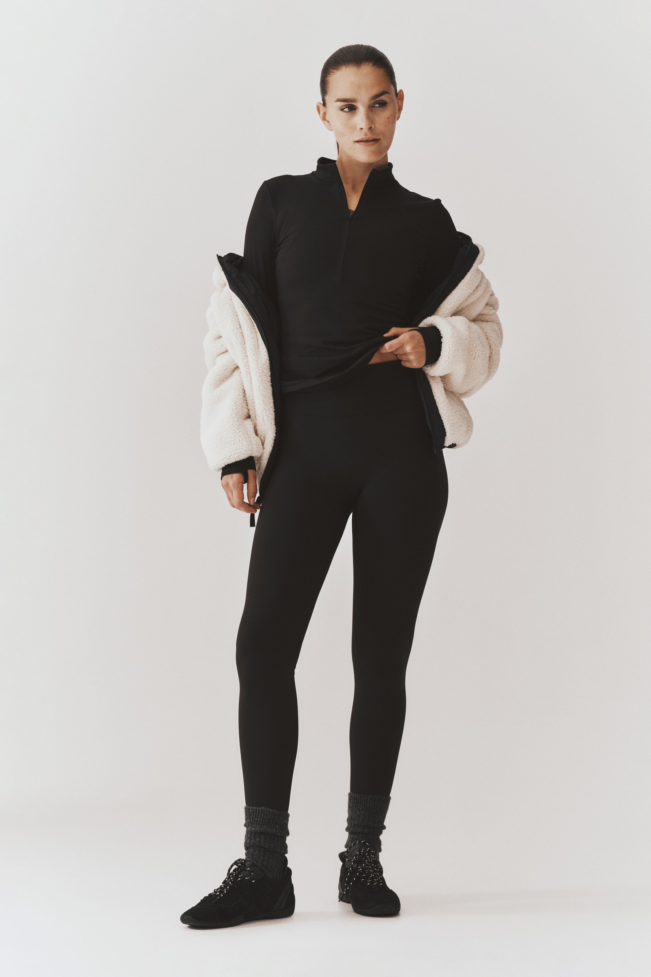Visa större bild: Modellen bär en svart långärmad topp med halv dragkedja och matchande leggings med hög midja, samtidigt som hon lyfter fållen på toppen med ena handen för att visa midjelinningen. En benvit fleecejacka bärs öppen ovanpå och stylingen kompletteras med mörkgrå ribbade strumpor och svarta sneakers.