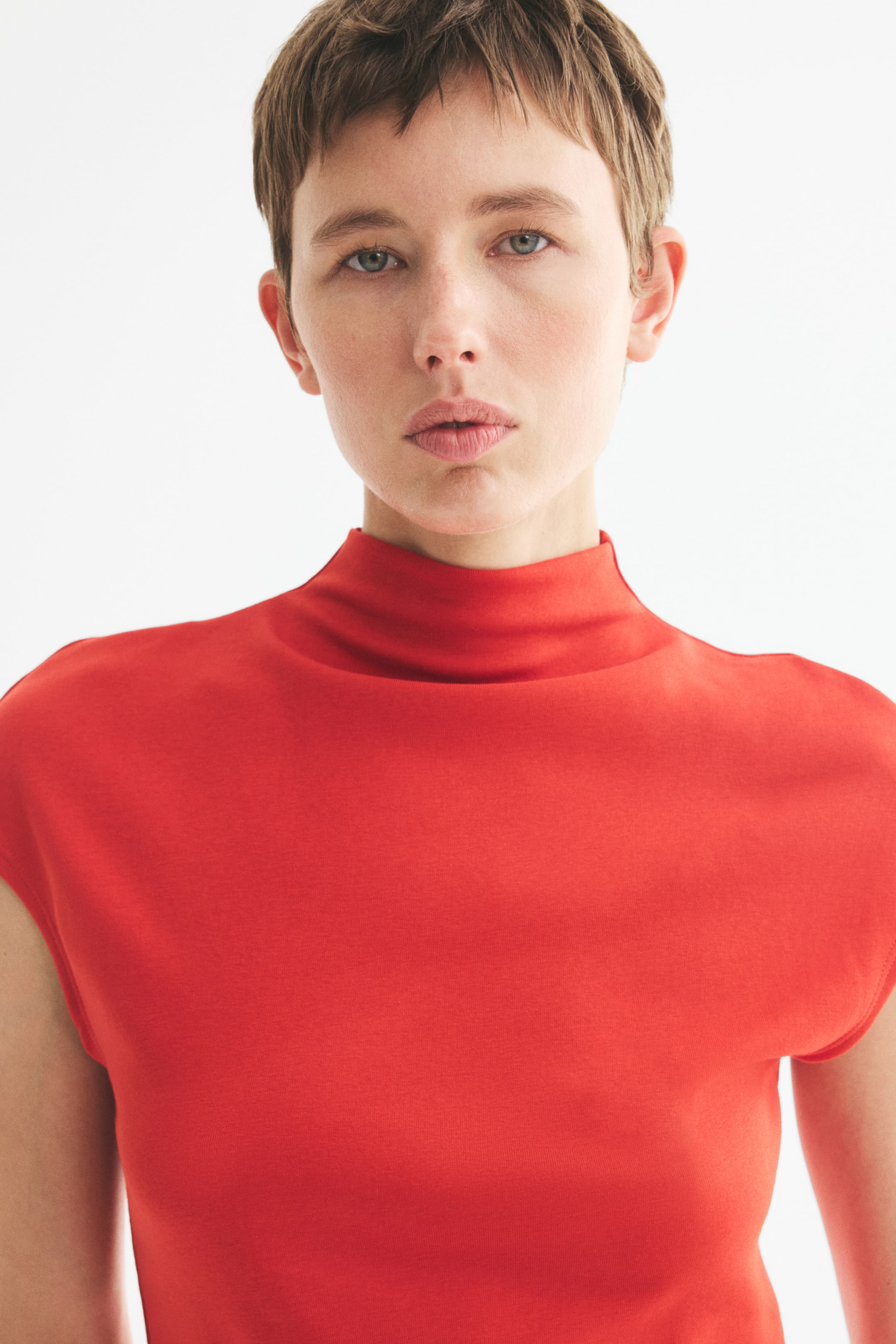 View larger image: The model is wearing a red fitted top made from soft cotton jersey, featuring a stand collar and cap sleeves. The top hugs the model's upper body closely.