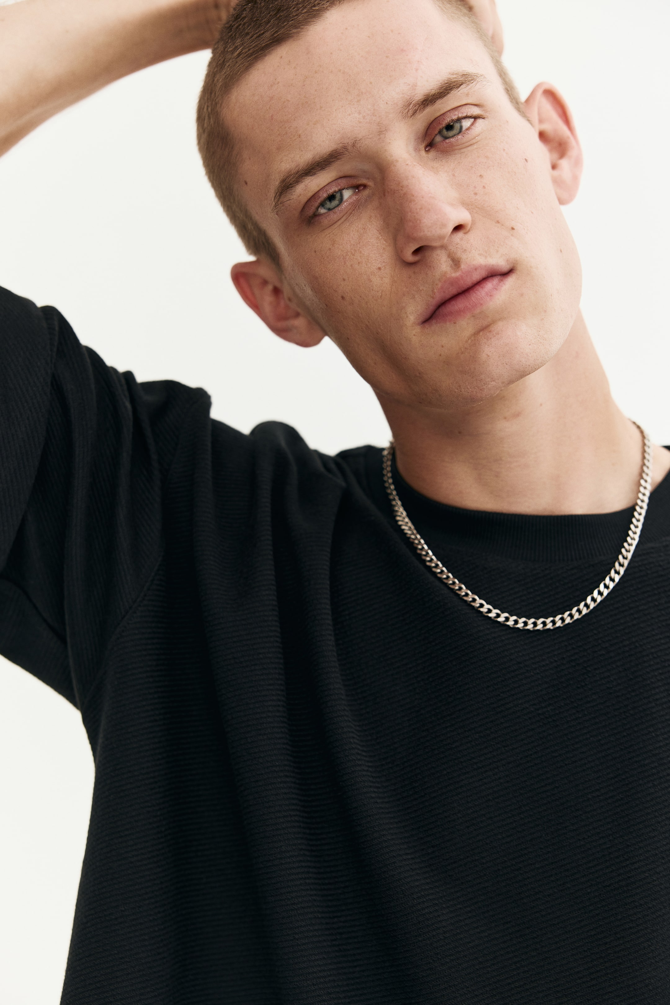 View larger image: A black short-sleeved top, featuring a crew neck and a subtle horizontally ribbed texture, is being worn with a silver chain necklace resting over its fabric.