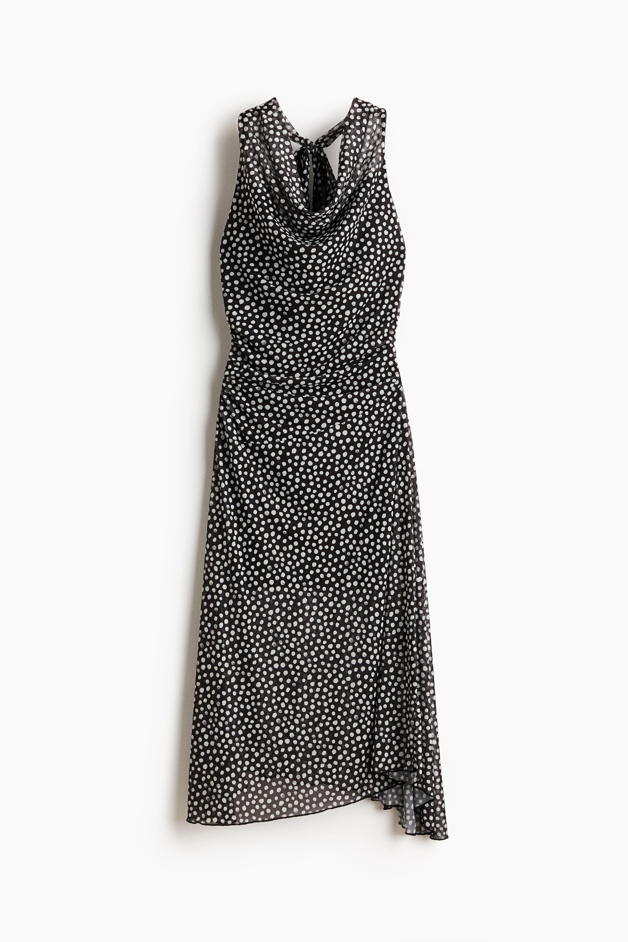 Ver imagen más grande: Un vestido negro con lunares blancos se muestra de frente, formando un escote drapeado (cowl) y un diseño sin mangas. Presenta tirantes halter en la nuca, fruncido en la zona media y una falda fluida con dobladillo asimétrico.
