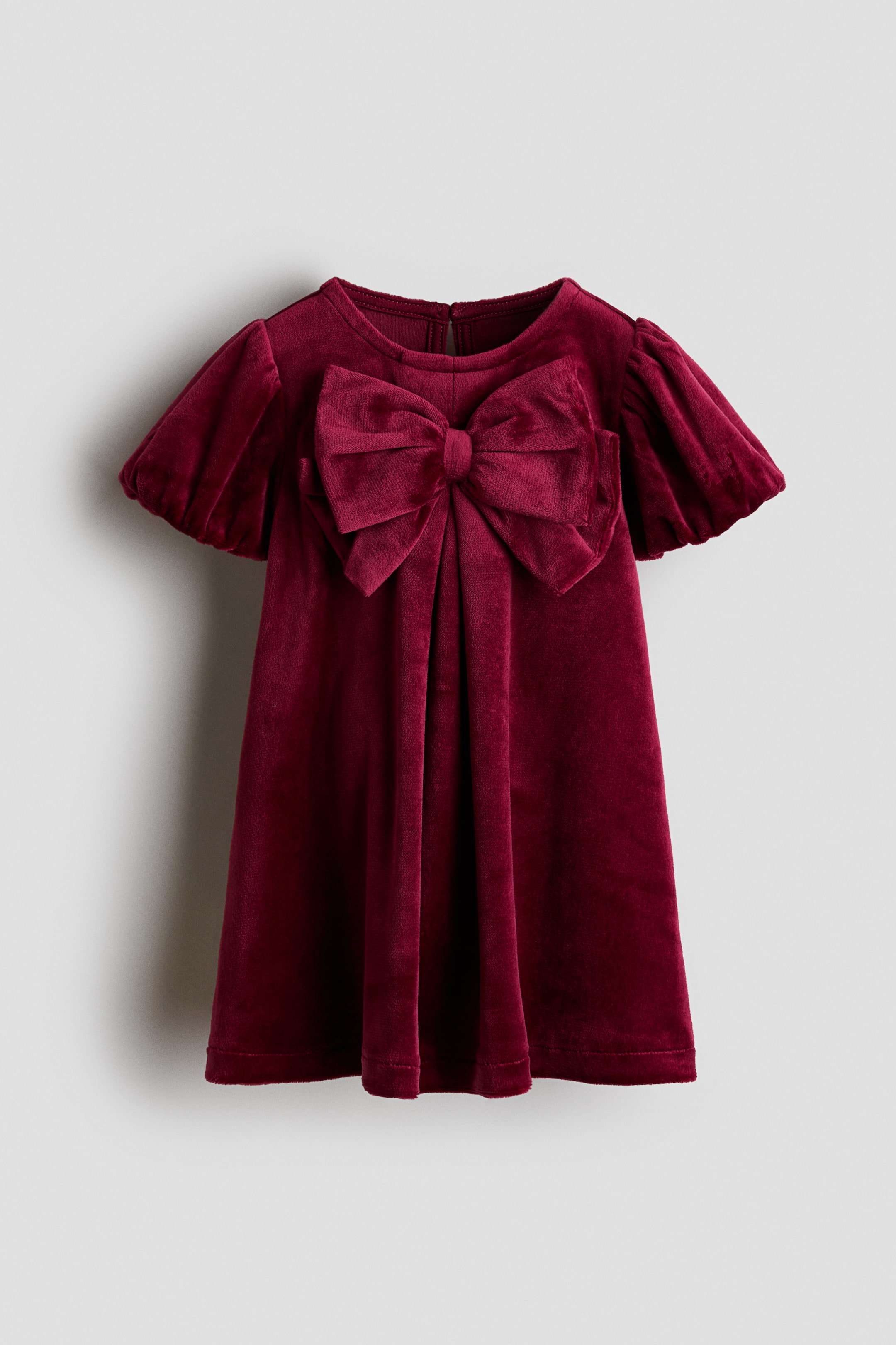 View larger image: A deep red velvet dress is shown from the front, featuring a large matching bow at the centre chest, short puff sleeves, and a crew neckline. The dress falls in a soft A-line silhouette with gentle pleats cascading from the base of the bow.