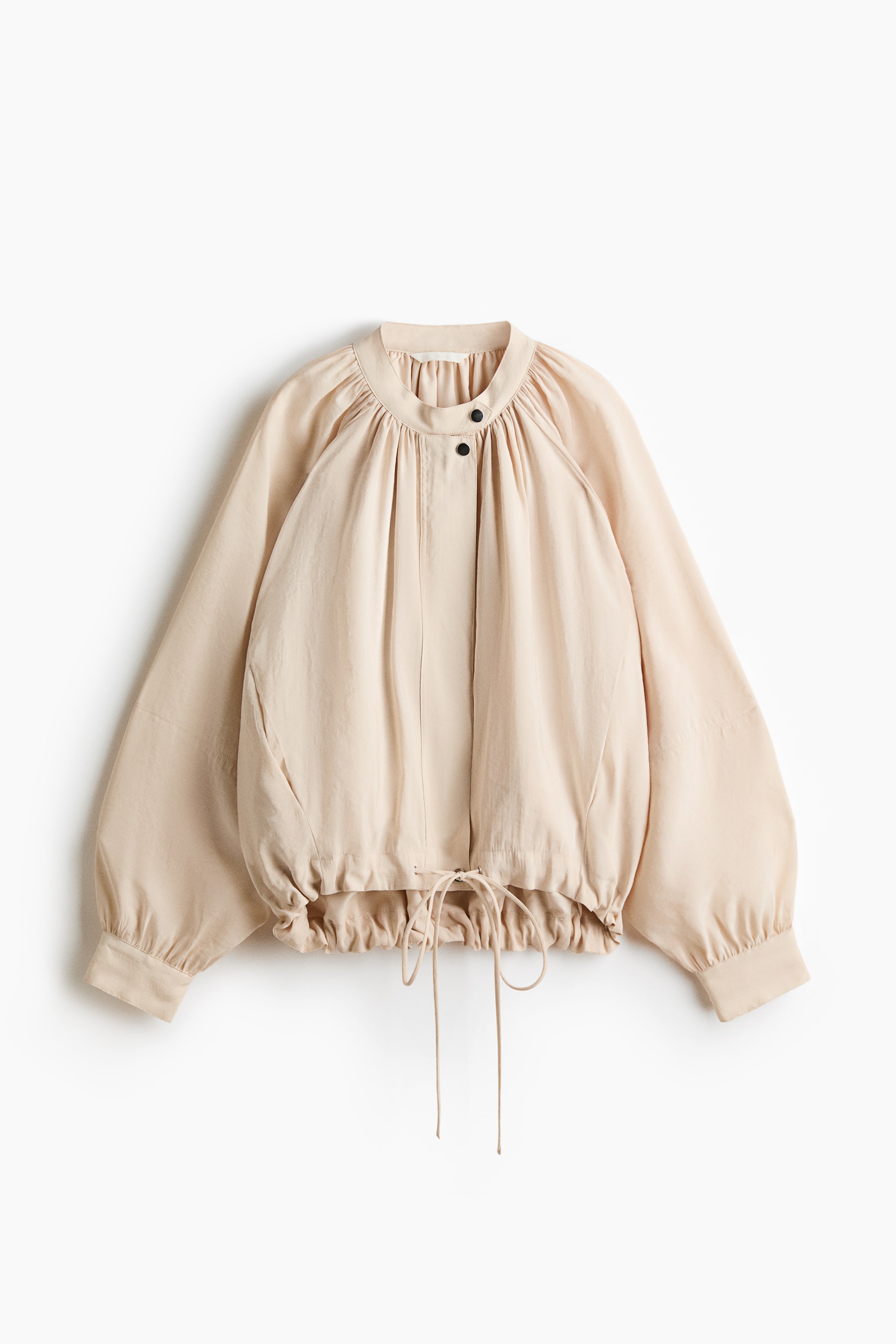 View larger image: An oversized light beige jacket is displayed from the front, showcasing a gathered band collar, a front placket fastened by two visible black press-studs, long raglan balloon sleeves with press-studs at the cuffs, and an elasticized hem with a visible drawstring.
