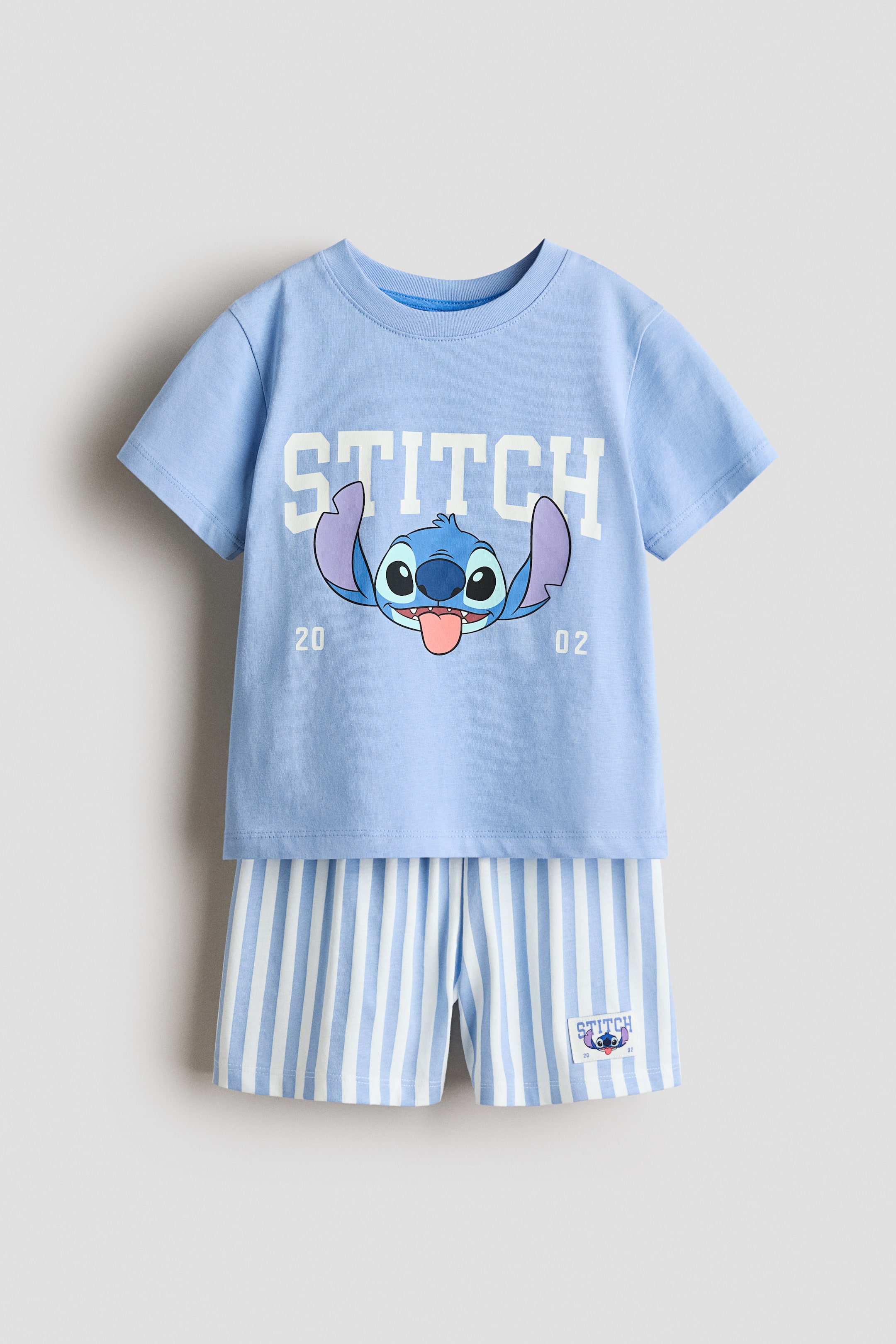 View larger image: A pair of pyjamas, crafted from soft, printed cotton jersey, is displayed flat and facing front. The light blue T-shirt showcases a ribbed trim around the neckline and a front print of the word "STITCH" along with the Stitch character and the numbers "20 02." The coordinating shorts are styled with white and light blue vertical stripes, an elasticated waist, and a small "STITCH" label.