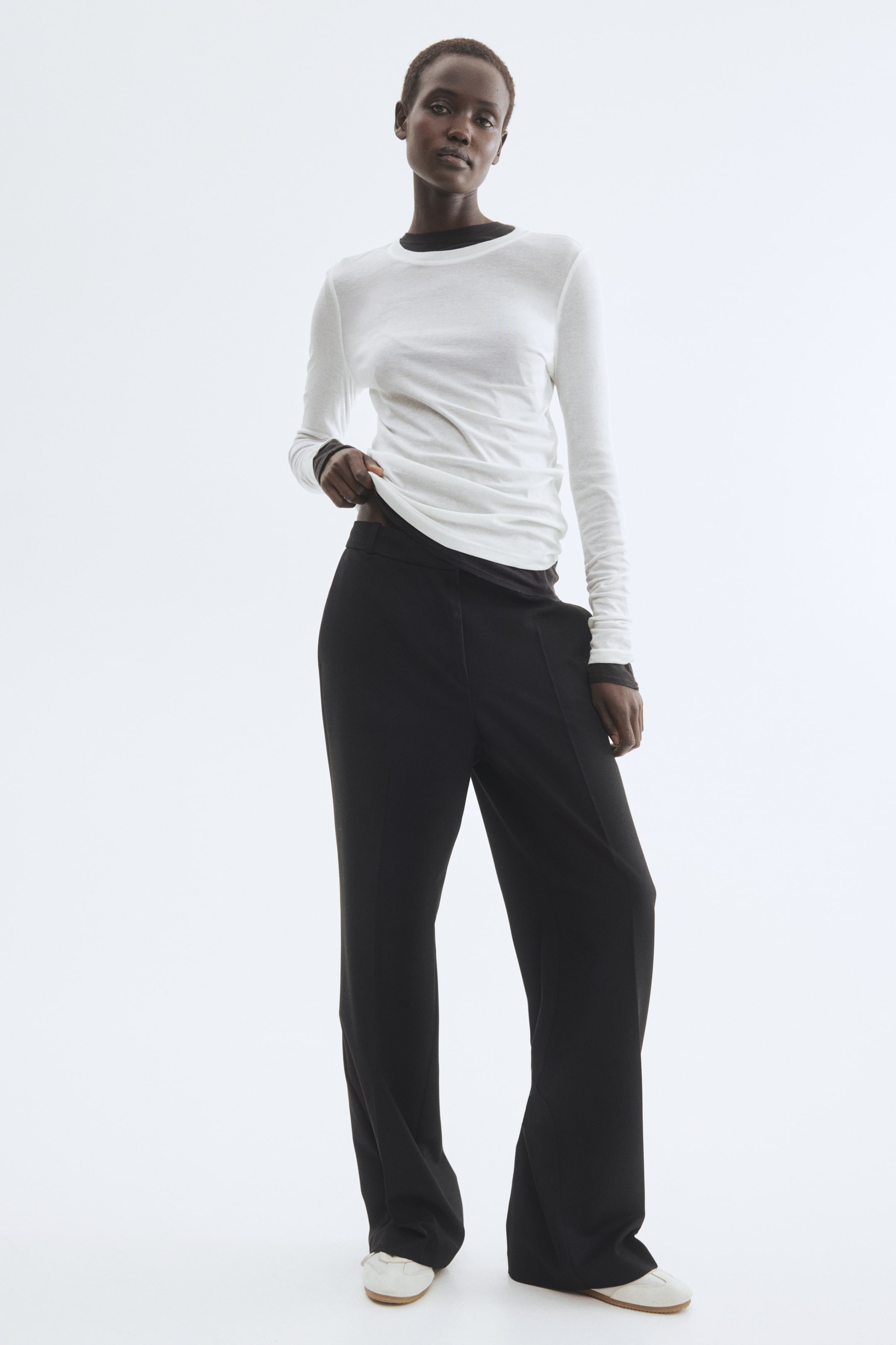 View larger image: Black tailored trousers featuring a regular waist, diagonal side pockets, and straight legs with front creases are worn by the model. The model is standing and adjusting her white top, revealing the waistband of the trousers.