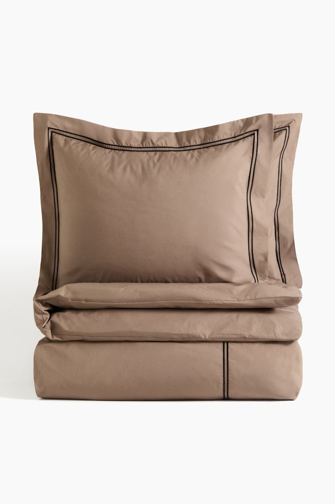A double duvet cover set is displayed, showcasing two pillowcases stacked atop a folded duvet cover. The light brown set features black double-line embroidery along the borders of the pillowcases and a matching vertical embroidery on the folded duvet cover. - 1