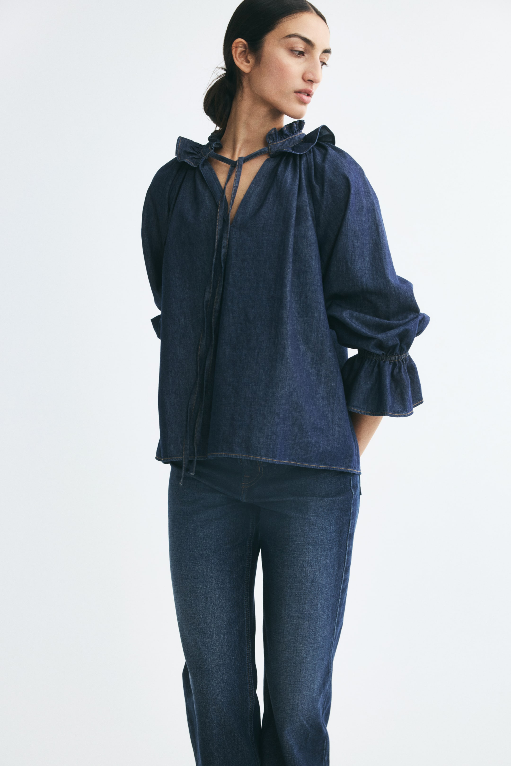 View larger image: A dark blue cotton denim blouse is being worn by the model, featuring a frilled collar with a V-shaped front opening and long, thin ties. The blouse has loose-fitting raglan sleeves with elasticated wrists and flounced cuffs, and is styled with matching blue denim jeans.