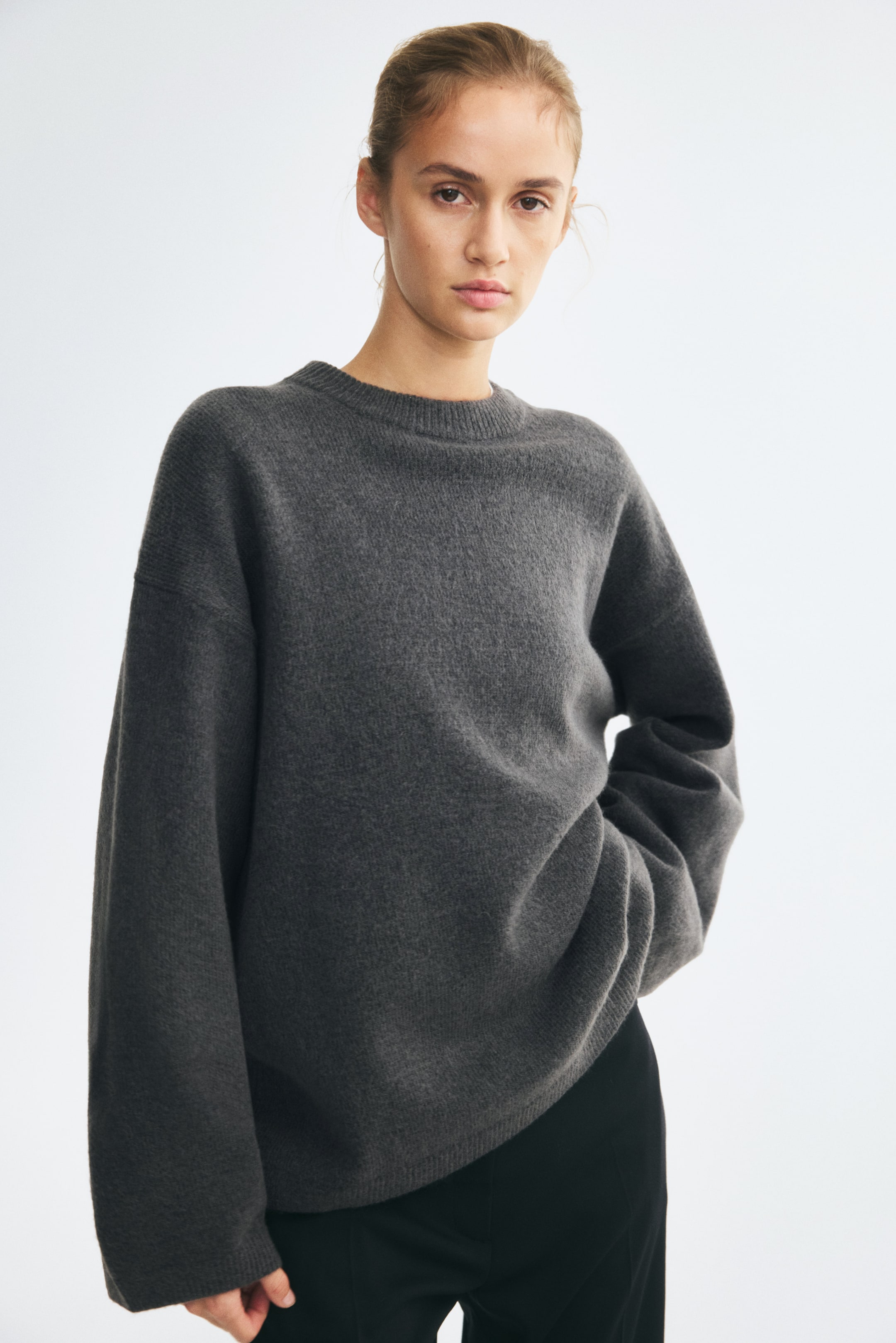 Ver imagen más grande: La modelo lleva puesto un jersey de punto gris oscuro de corte oversized, con cuello redondo acanalado, hombros caídos y mangas largas. Posea con una mano metida en el bolsillo del pantalón negro, haciendo que el bajo acanalado del jersey caiga y se frunza ligeramente en el lateral.