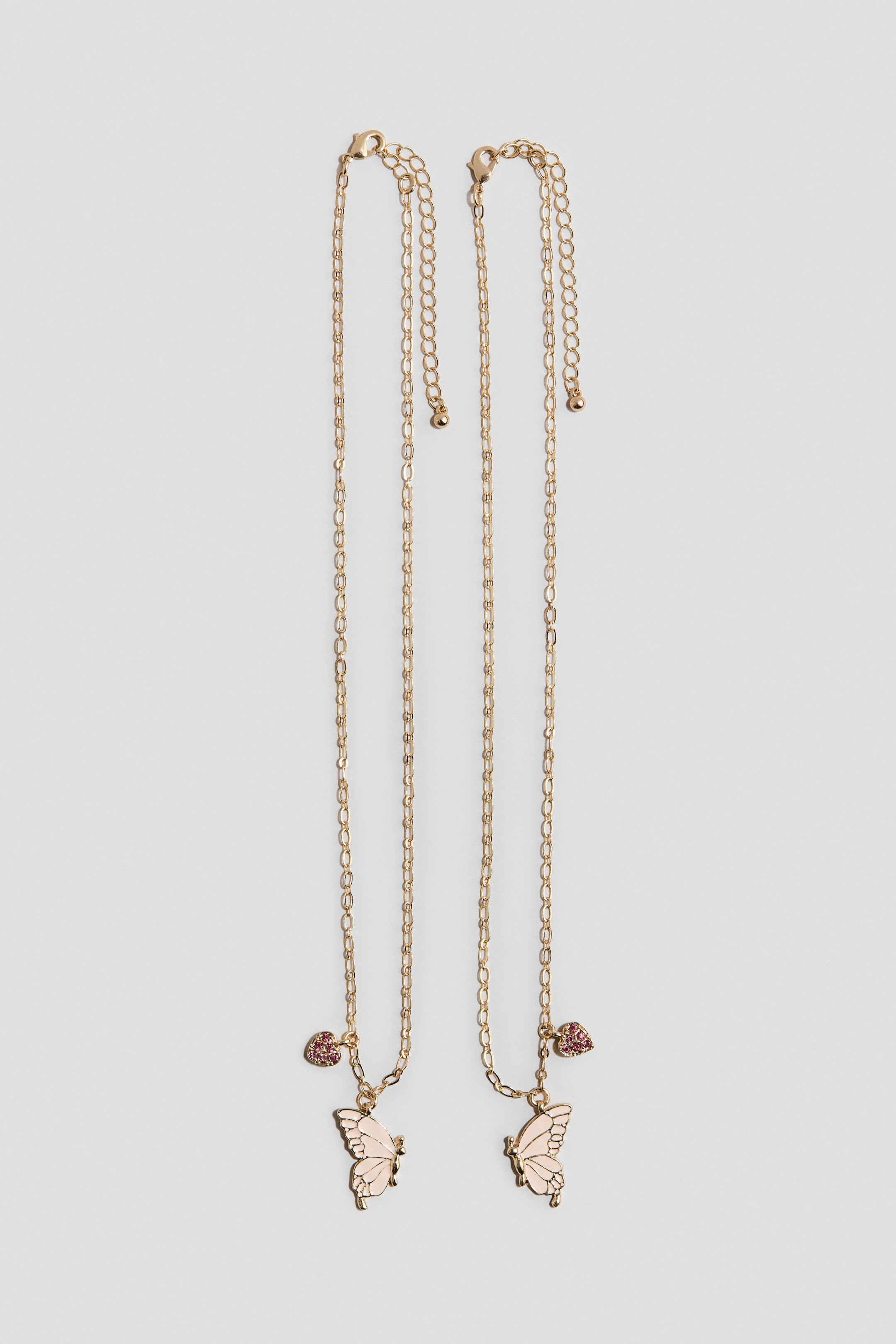 Grotere afbeelding bekijken: Twee goudkleurige metalen kettinghalskettingen hangen verticaal, elk voorzien van een verstelbare verlengkettinkje met een karabijnsluiting. Elke ketting toont een lichtroze geëmailleerde vlinderhanger en een kleinere met roze edelstenen bezette hartbedel, beide naar voren gericht.