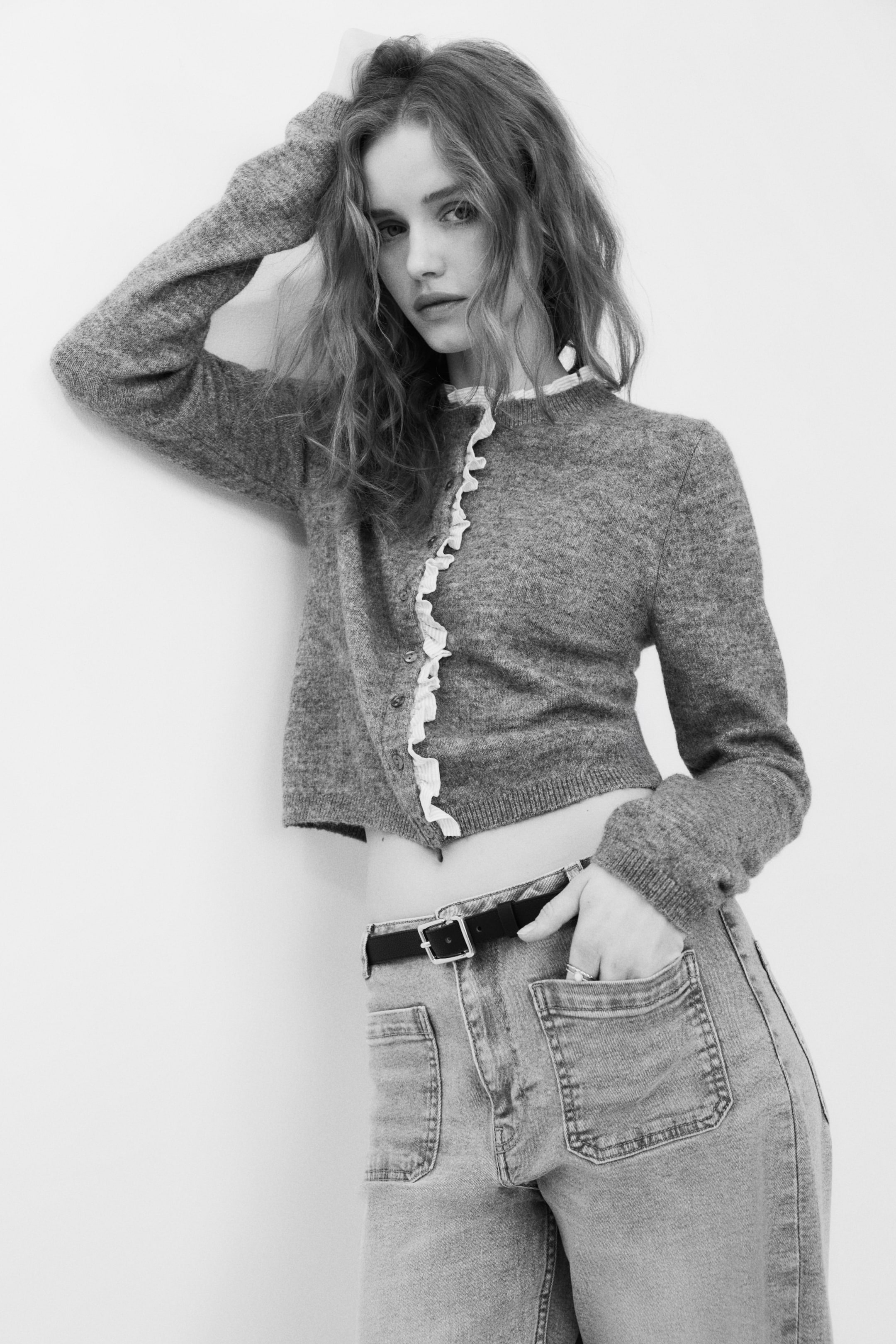 Ampliar la imagen: La modelo lleva puesto un cárdigan ajustado de tejido fino, que presenta un escote redondo con ribete acanalado y un detalle de volante en tono claro en el escote y la banda de botones, con botones que cierran la parte delantera. Tiene mangas largas con puños acanalados y un dobladillo acanalado, y está diseñado en un largo recortado, combinado con jeans.