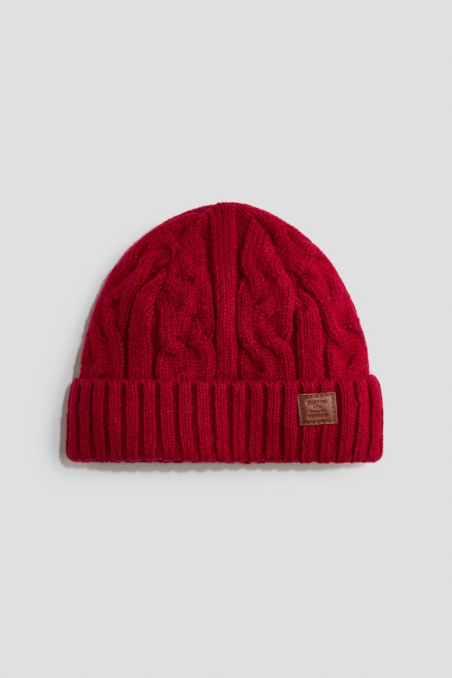 Un bonnet rouge en maille torsadée est présenté de face, avec un motif torsadé texturé sur la calotte et un large revers côtelé replié. Une petite étiquette rectangulaire en cuir marron est cousue sur le côté droit du revers. - 1