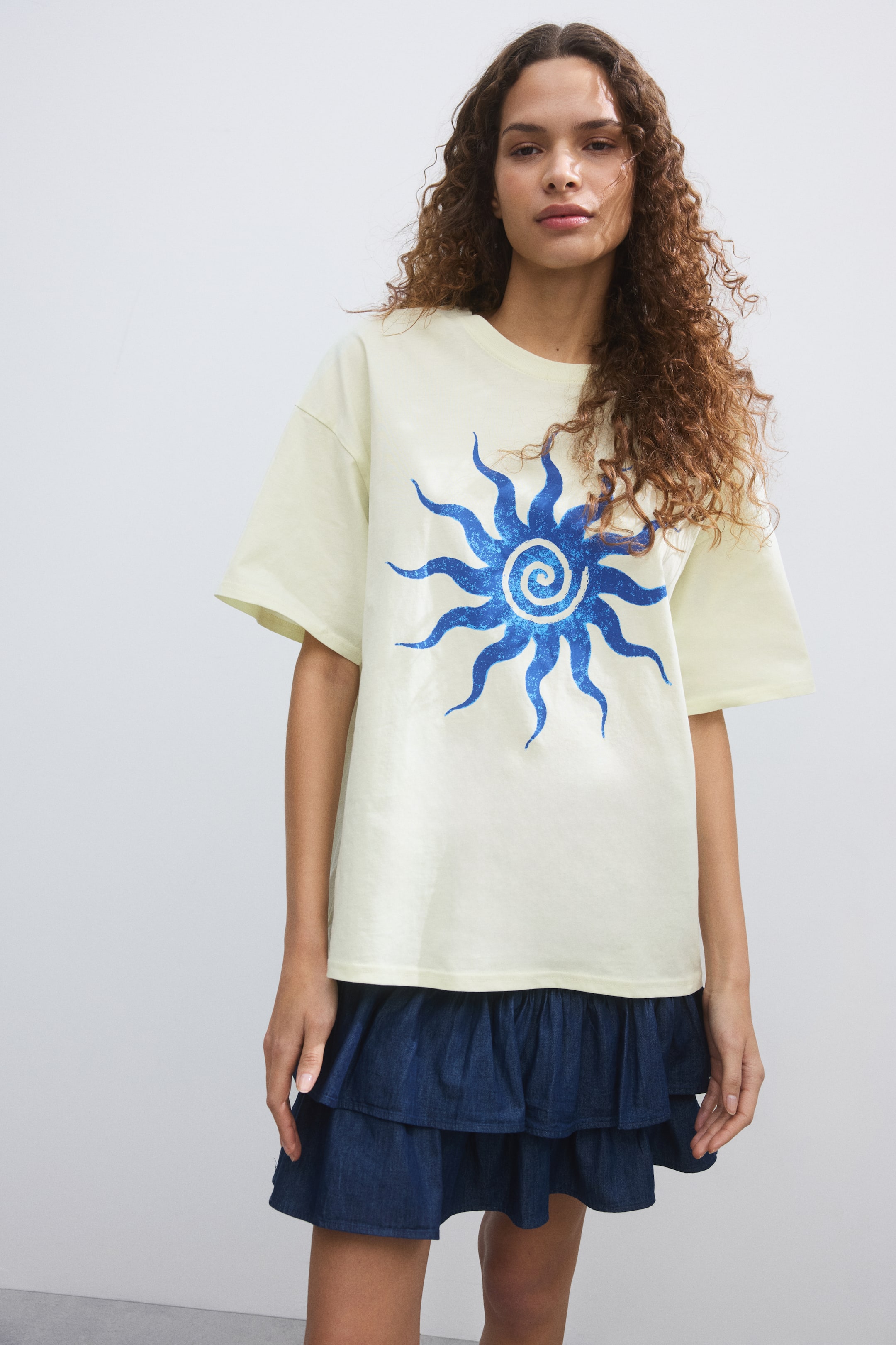 View larger image: The pale yellow t-shirt showcases a striking, shimmering blue sun graphic with a spiral center on its front. Designed with a crew neck and short, wide sleeves, it offers a relaxed fit.