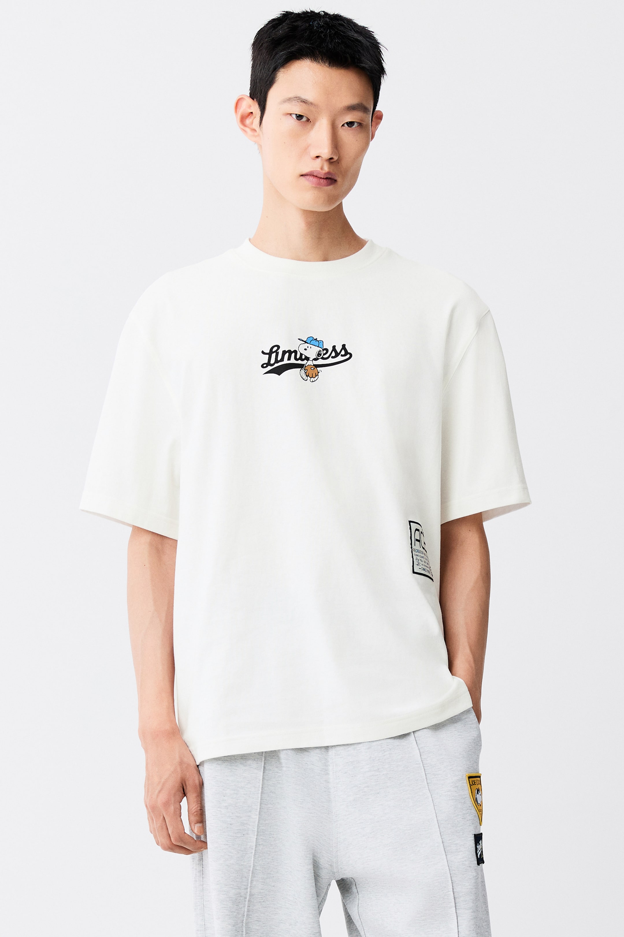 View larger image: A cream-coloured heavyweight cotton jersey T-shirt is worn by the model, featuring a loose fit, a ribbed crew neck, and gently dropped shoulders. The T-shirt features a printed graphic on the chest with the word "Limitless" alongside a cartoon character, and is paired with light grey sweatpants.