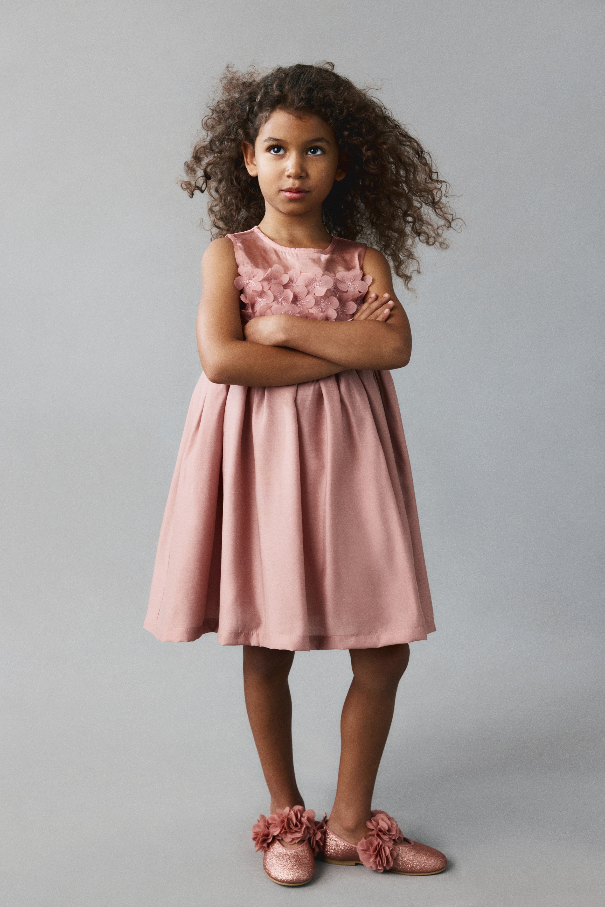 A dusty pink sleeveless dress is shown from the front, featuring a sheer mesh upper yoke and a round neckline. The empire waistline is decorated with rows of 3D fabric flower appliquÃ©s with embroidered details, leading into a gathered, flared skirt.