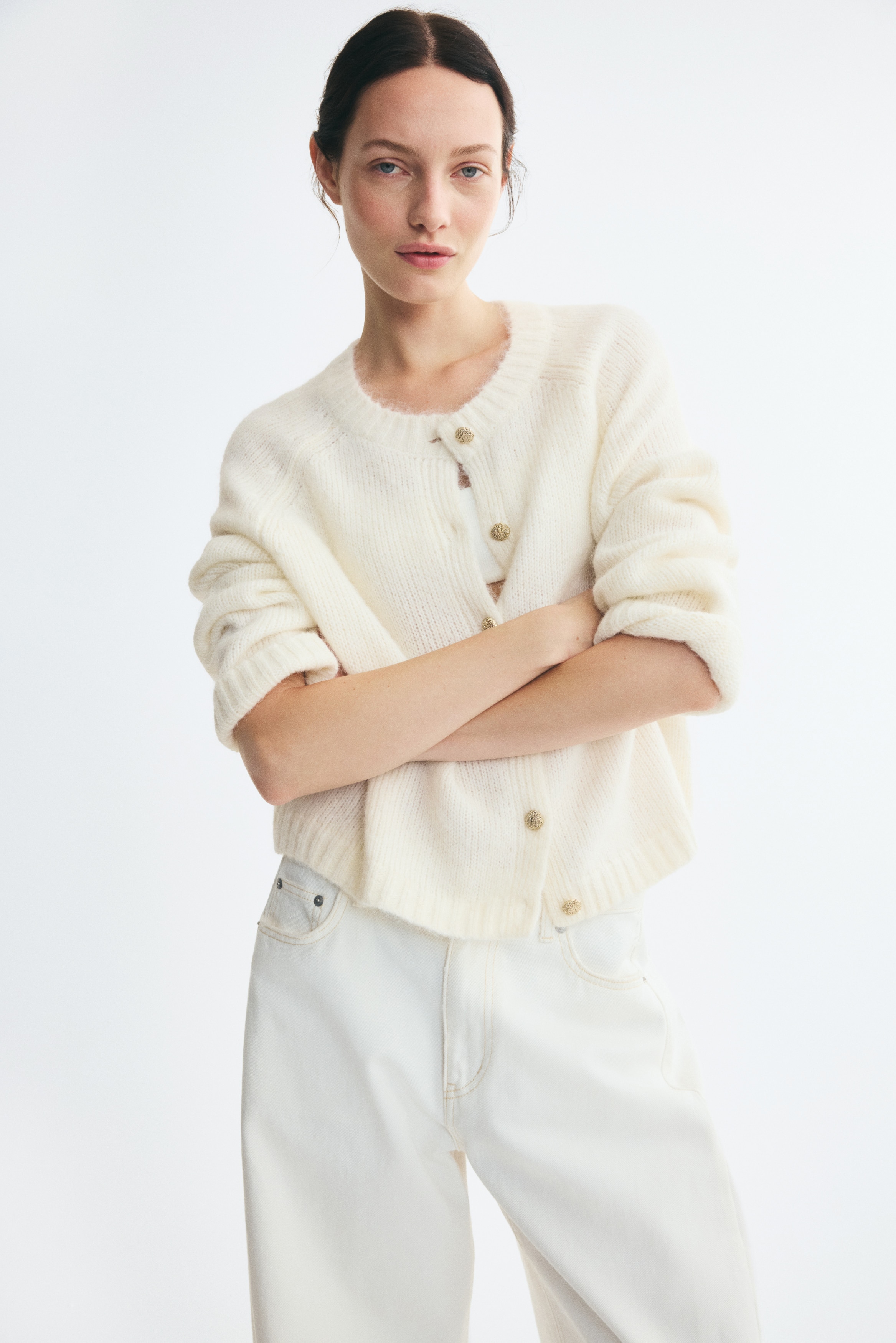 An off-white knit cardigan is shown facing front, showcasing a soft, fuzzy texture and a round, ribbed neckline. It features a front button closure with six decorative gold-toned buttons, long sleeves with ribbed cuffs, and a ribbed hem.