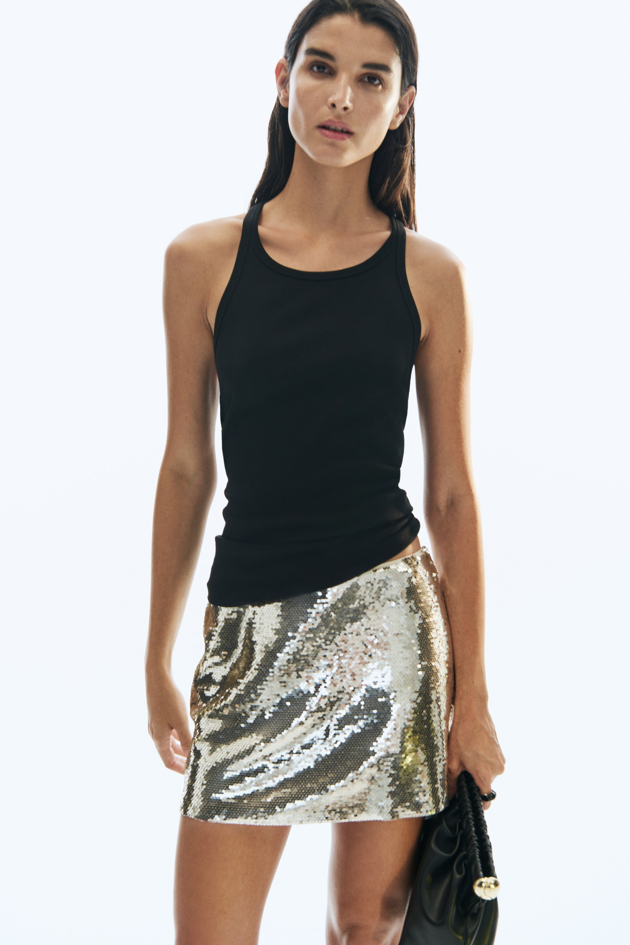 View larger image: A sequinned mesh mini skirt featuring shimmering silver sequins is being worn by the model. The skirt is styled with a fitted black tank top layered over the waistband, and a black handbag is being held at the side.