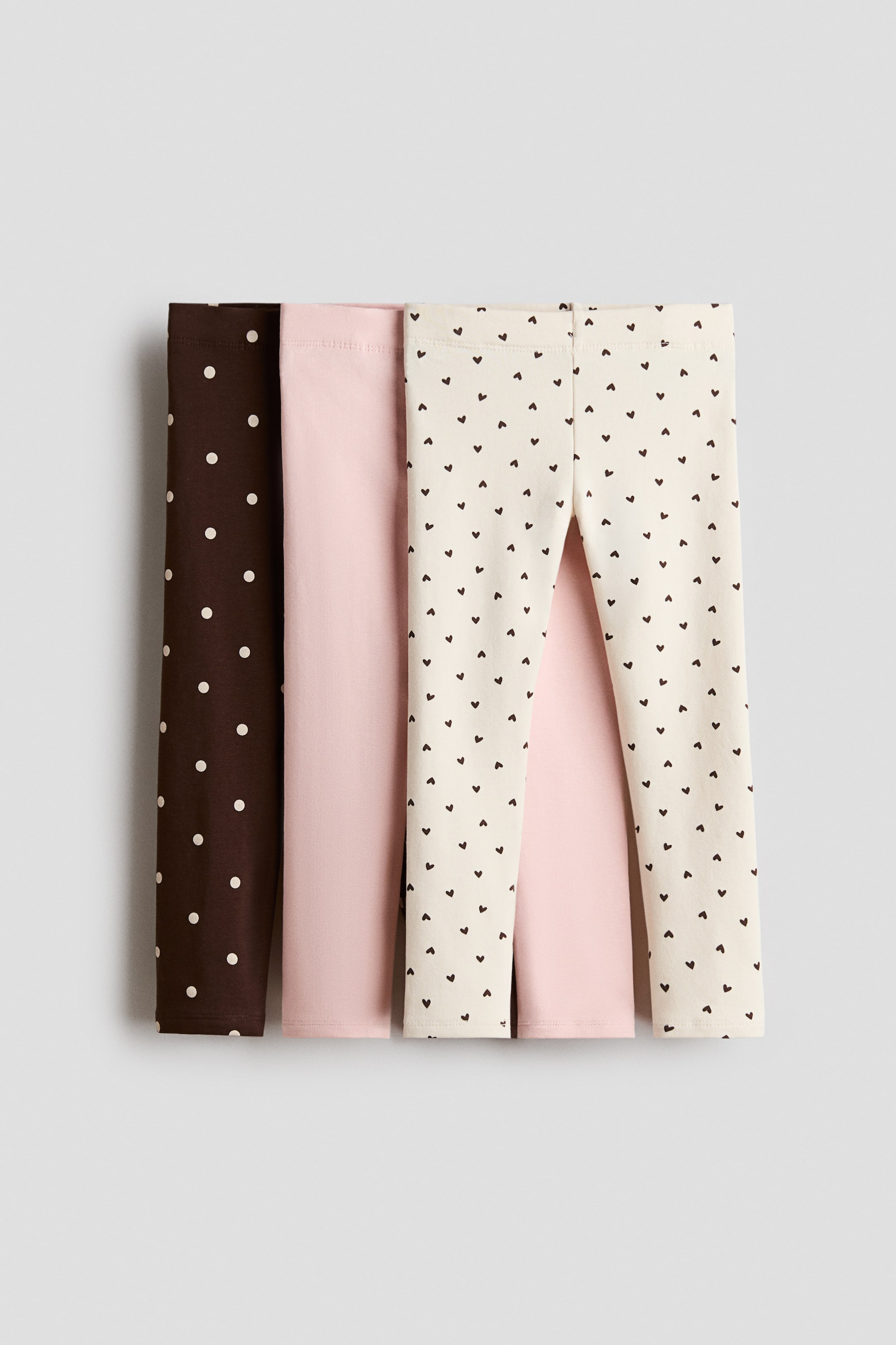 View larger image: Three pairs of lightweight leggings are displayed front-facing in a layered arrangement. The selection includes an off-white pair featuring a small dark heart pattern in the foreground, a solid light pink pair in the middle, and a dark brown pair with white polka dots at the back, all showcasing elasticated waistbands and a slim-fit design.