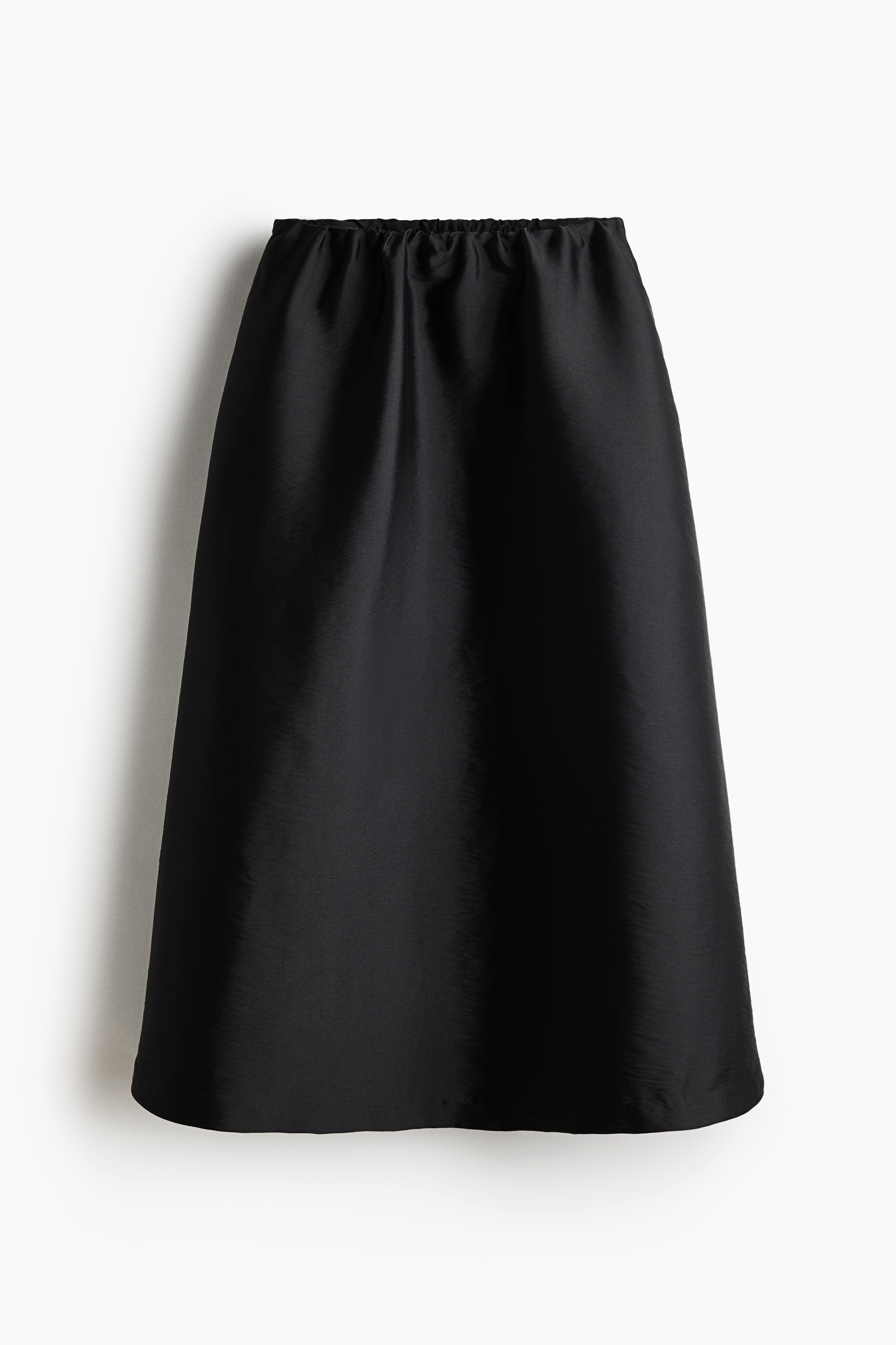 Une jupe noire trapÃ¨ze, longueur mi-mollet, avec un lÃ©ger aspect brillant et une ceinture froncÃ©e est portÃ©e par le mannequin, qui se tient debout, les mains dans les poches latÃ©rales. La jupe est associÃ©e Ã  un haut noir ajustÃ© Ã  manches longues et Ã  des sandales noires Ã  brides.