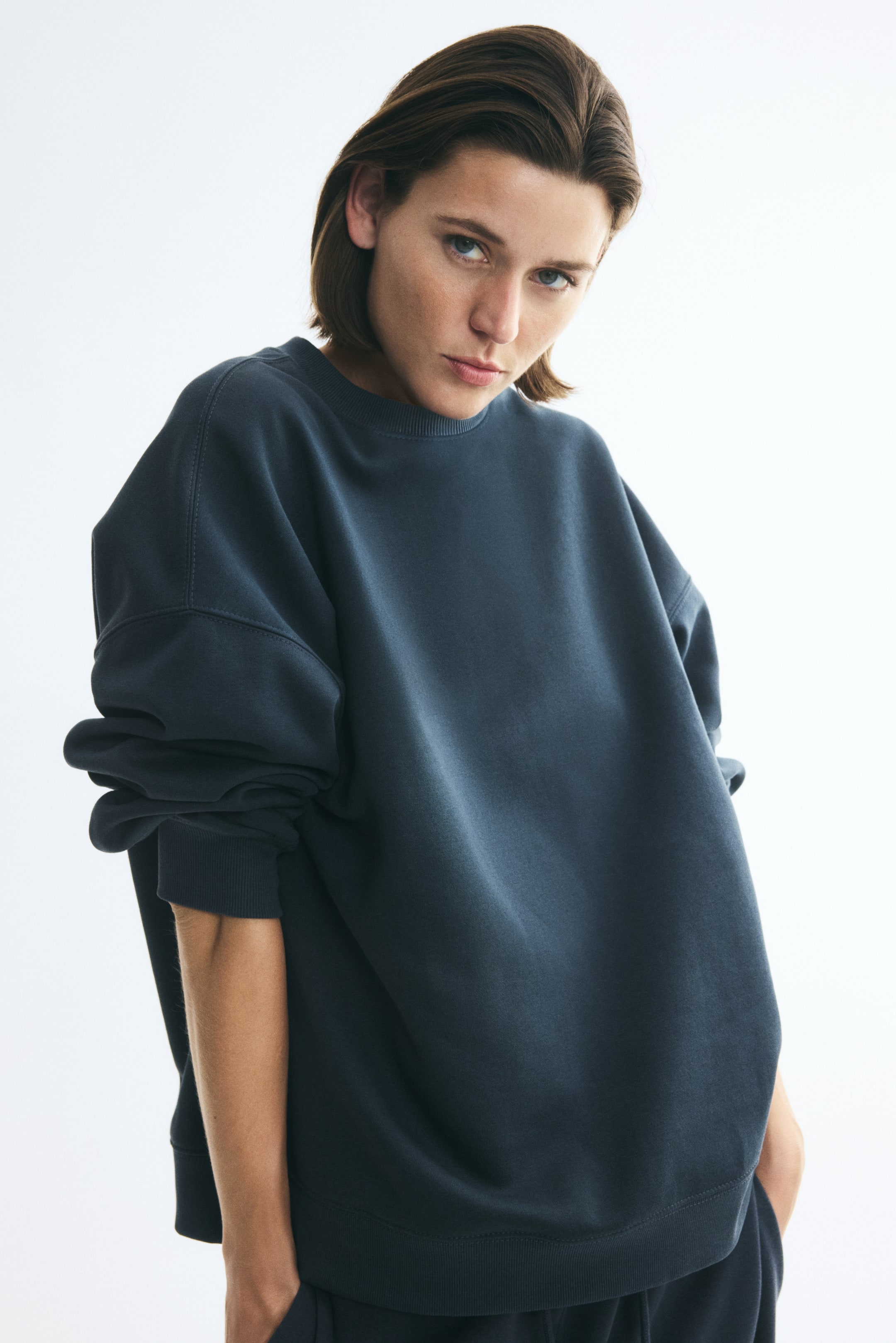 View larger image: The model is wearing an oversized dark gray-blue sweatshirt with a round ribbed neckline, dropped shoulders, and ribbed cuffs and hem. The model is standing with their hands in the pockets of matching dark trousers.