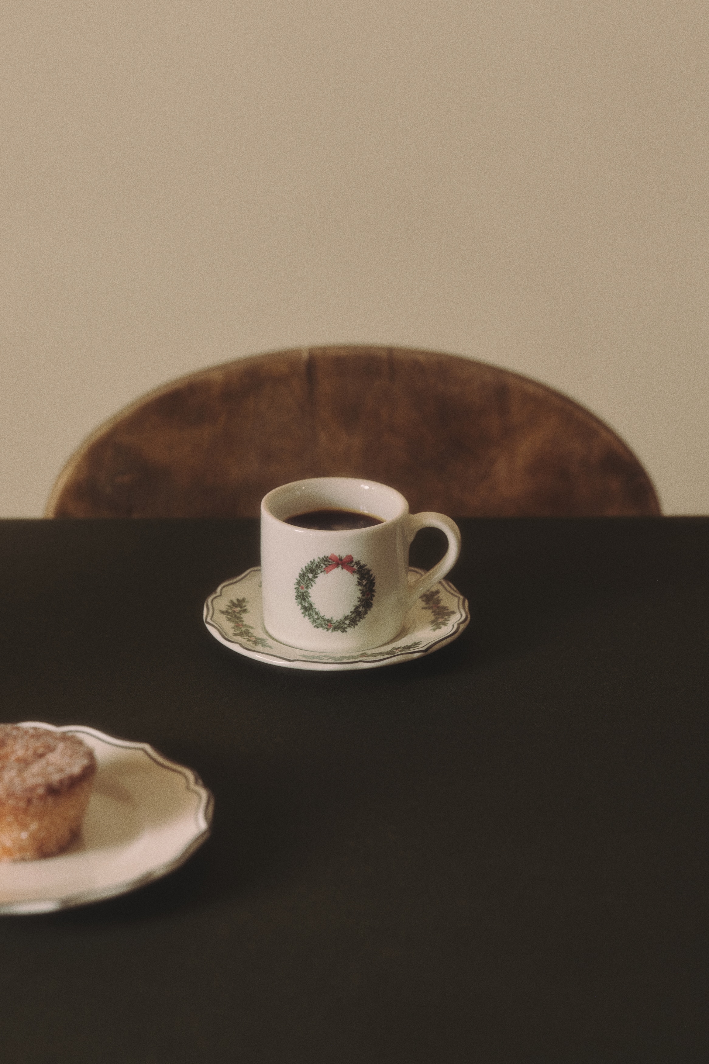 Two white ceramic cups rest on matching scalloped saucers in a front-facing view, with each cup displaying a green wreath illustration with a small red bow. The saucers feature a delicate, dark-rimmed wavy edge.