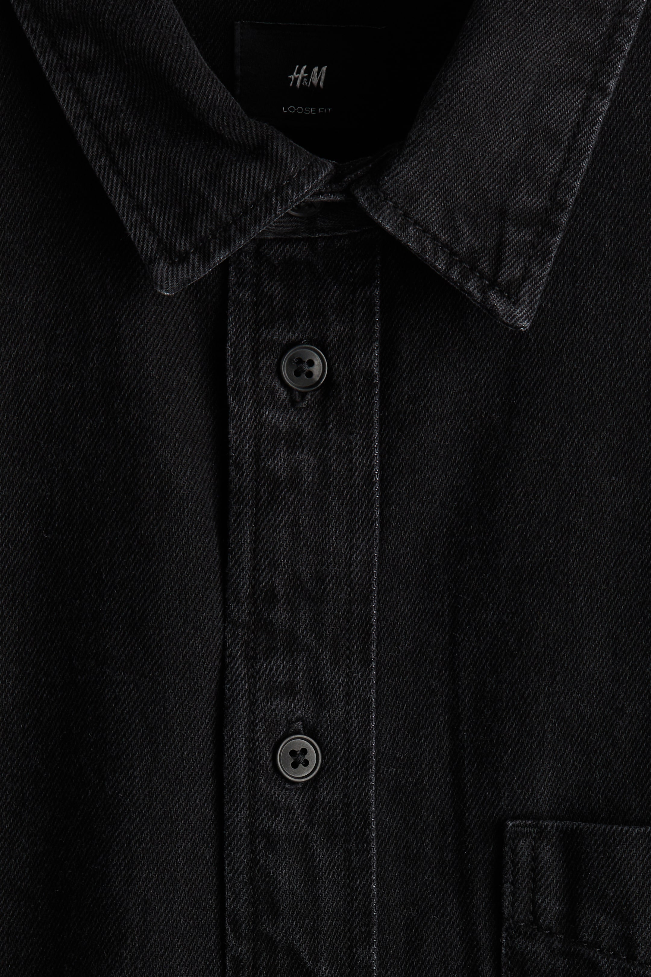 Agrandir l'image: Une chemise noire en sergé est exposée de FACE, avec un col pointu et une patte de boutonnage avec des boutons à quatre trous gris foncé. Une poche poitrine est visible à droite, et une étiquette noire avec "H&M" et "LOOSE FIT" est visible sous le col.