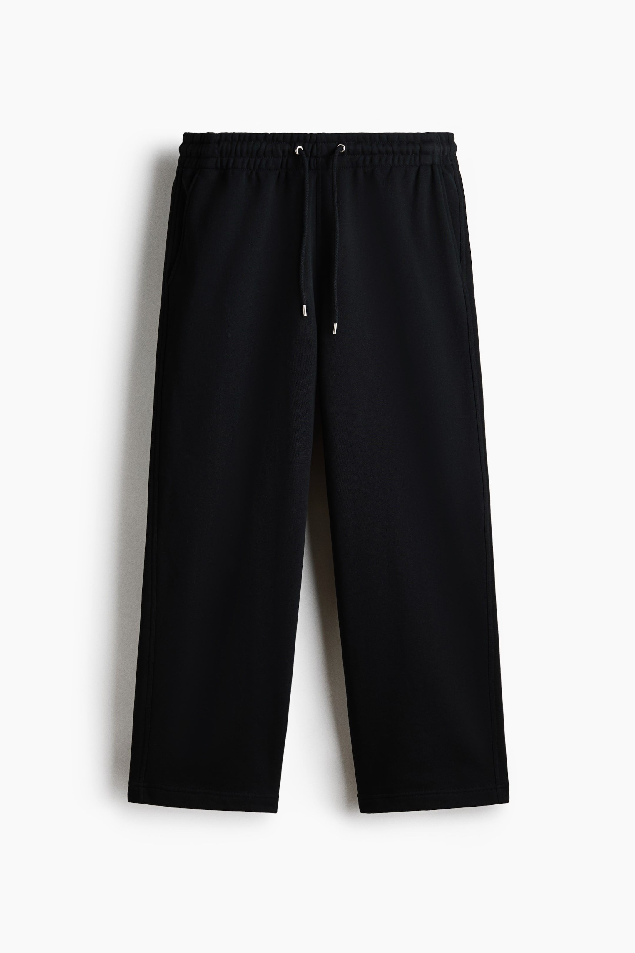 拡大画像を表示: コットンスウェット生地のブラックスウェットパンツが正面から見た様子を示しています。カバーされたゴムウエストにはドローストリングが付いており、銀色のメタルチップで終わっています。また、サイドポケットがあり、ストレートレッグのルーズフィットを特徴としています。