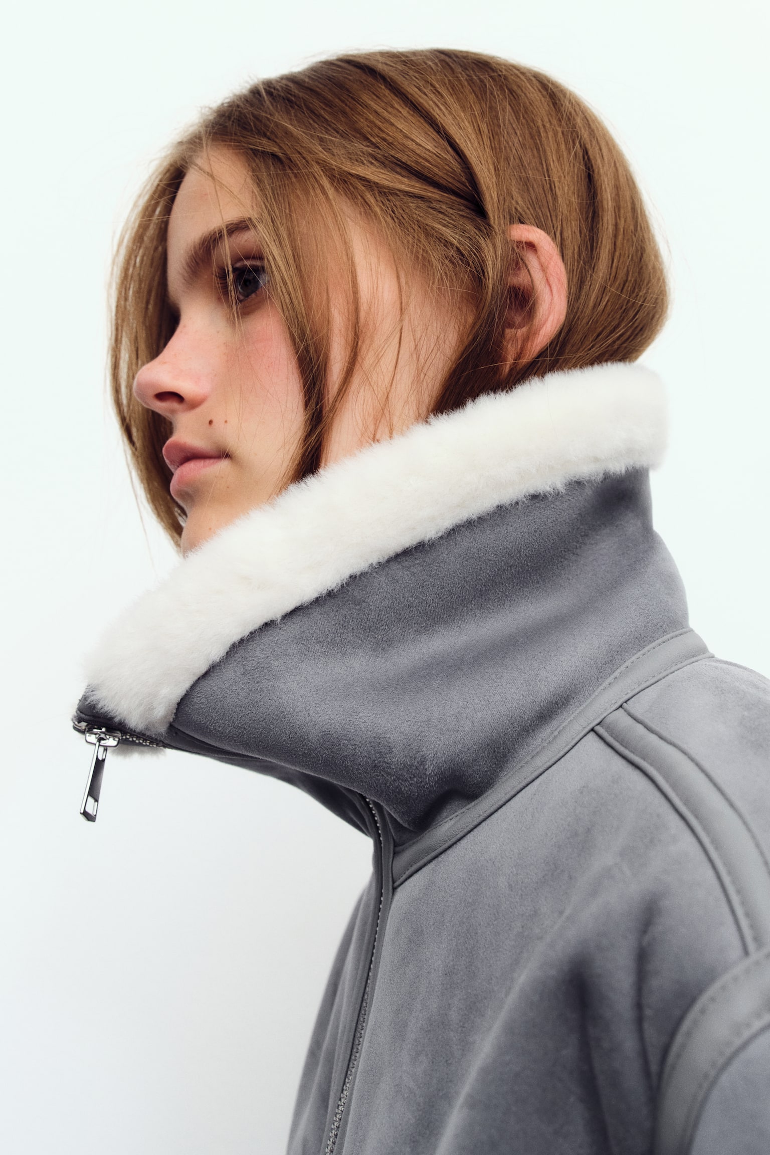 A grey cropped jacket is shown front-on, featuring a wide white faux shearling collar, cuffs and hem. A full-length zip runs down the front, and side welt pockets are visible. - 1