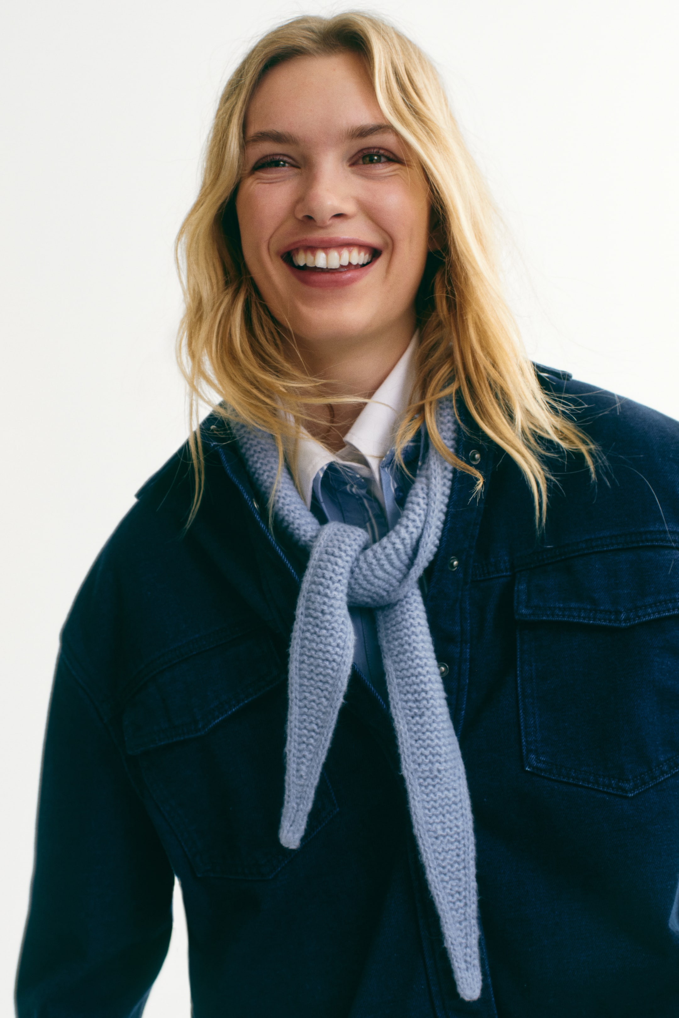 View larger image: A light-blue scarf in a soft garter-stitch knit with pointed ends is worn by the model. It's tied around her neck, styled over a white shirt and under a dark-blue jacket.