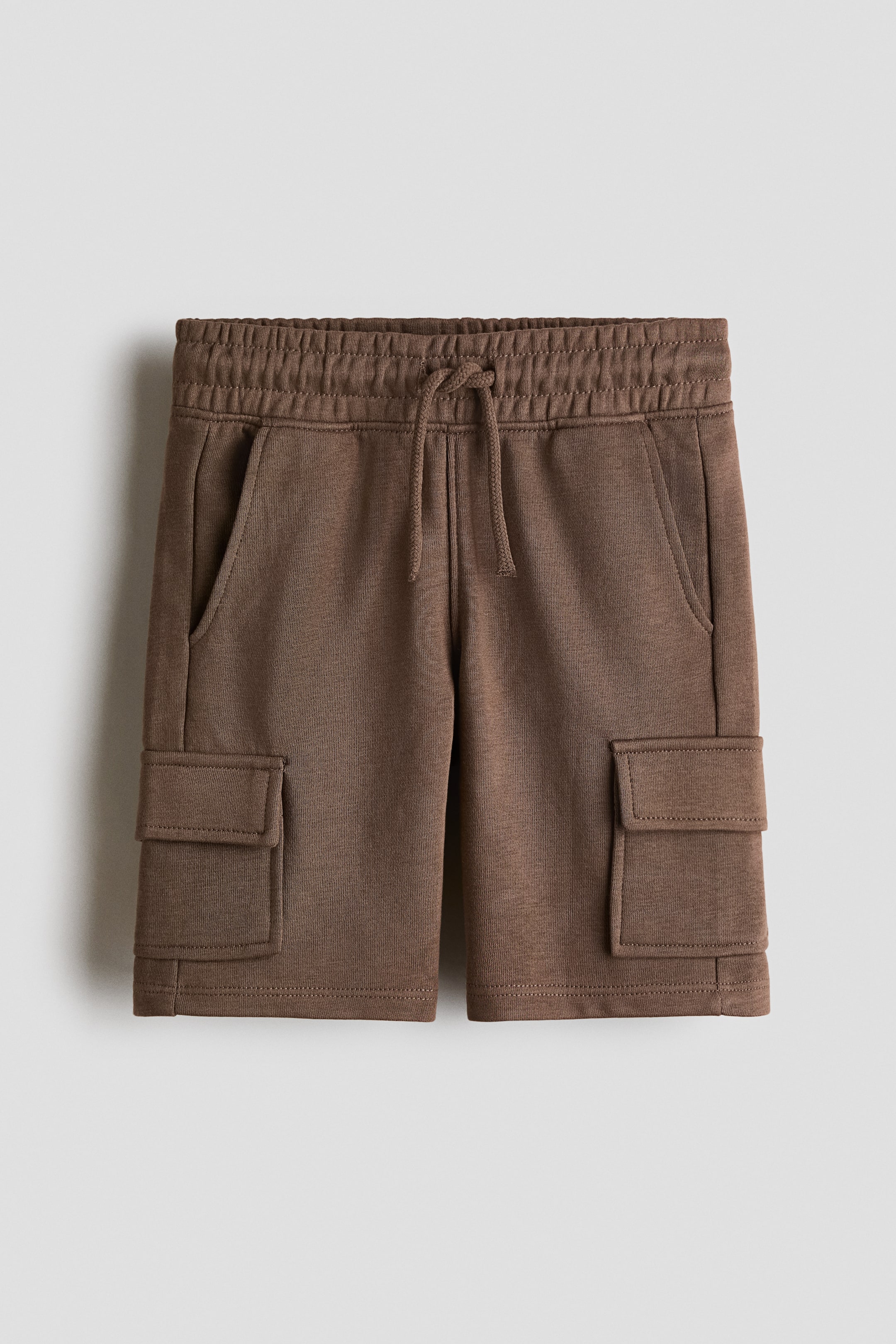 View larger image: Brown knee-length cargo shorts crafted from lightweight sweatshirt fabric are showcased from the front, featuring an elasticated, drawstring waist, diagonal side pockets, and flap leg pockets on each leg.