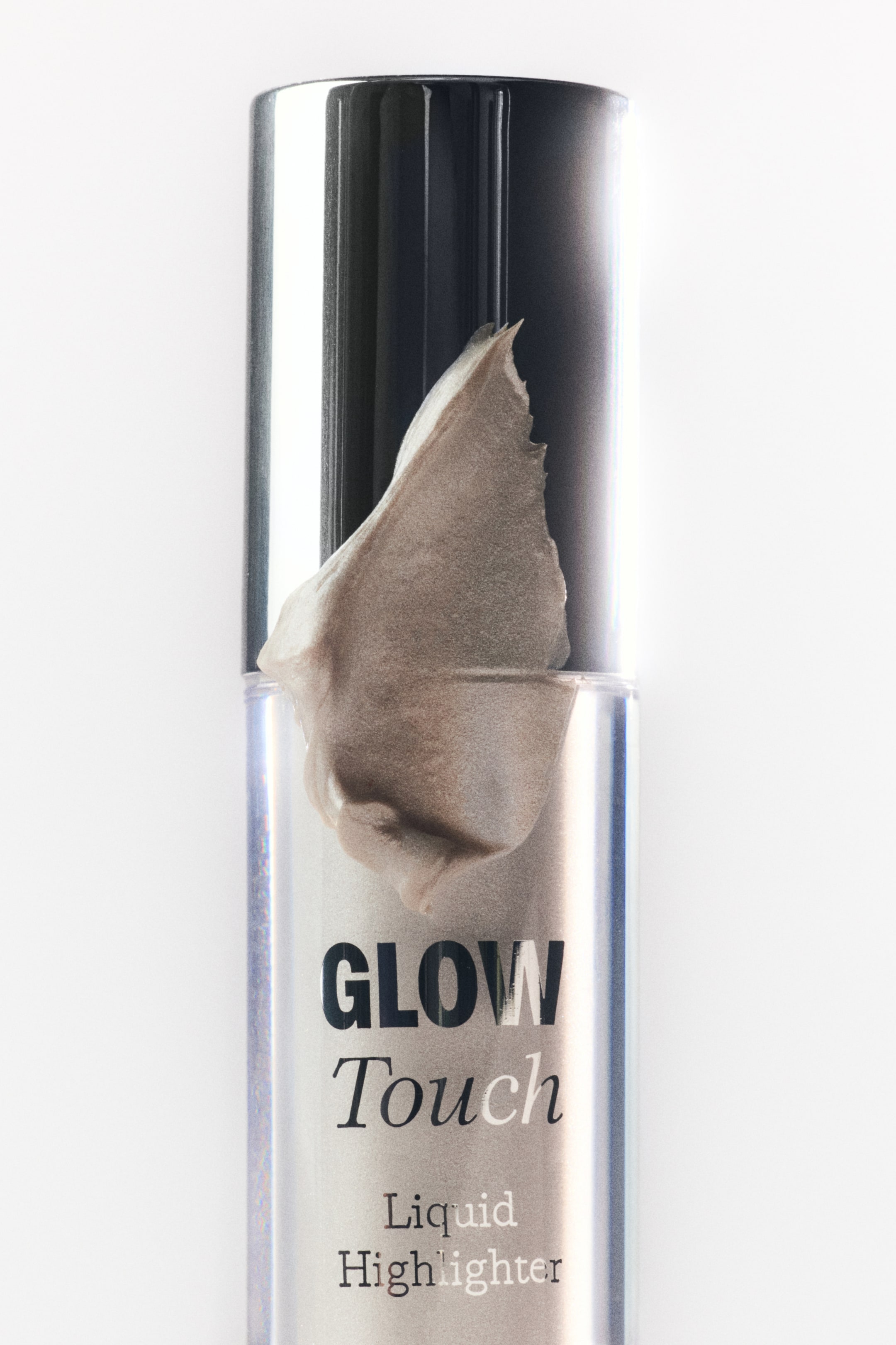 Visa större bild: Ett cylindriskt flytande highlighter-rör står upprätt, med en mörk reflekterande kork och en genomskinlig kropp. En skimrande, ljusbrons flytande highlighter är synligt applicerad på den övre kanten av det genomskinliga röret och korken. Texten "GLOW Touch Liquid Highlighter" är tryckt på röret.