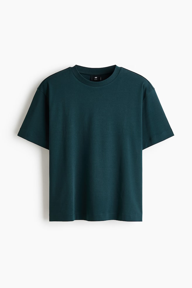 A dark teal T-shirt is shown from the front, featuring a loose, boxy fit with a round, rib-trimmed neckline, dropped shoulders, and a straight-cut hem. - 1