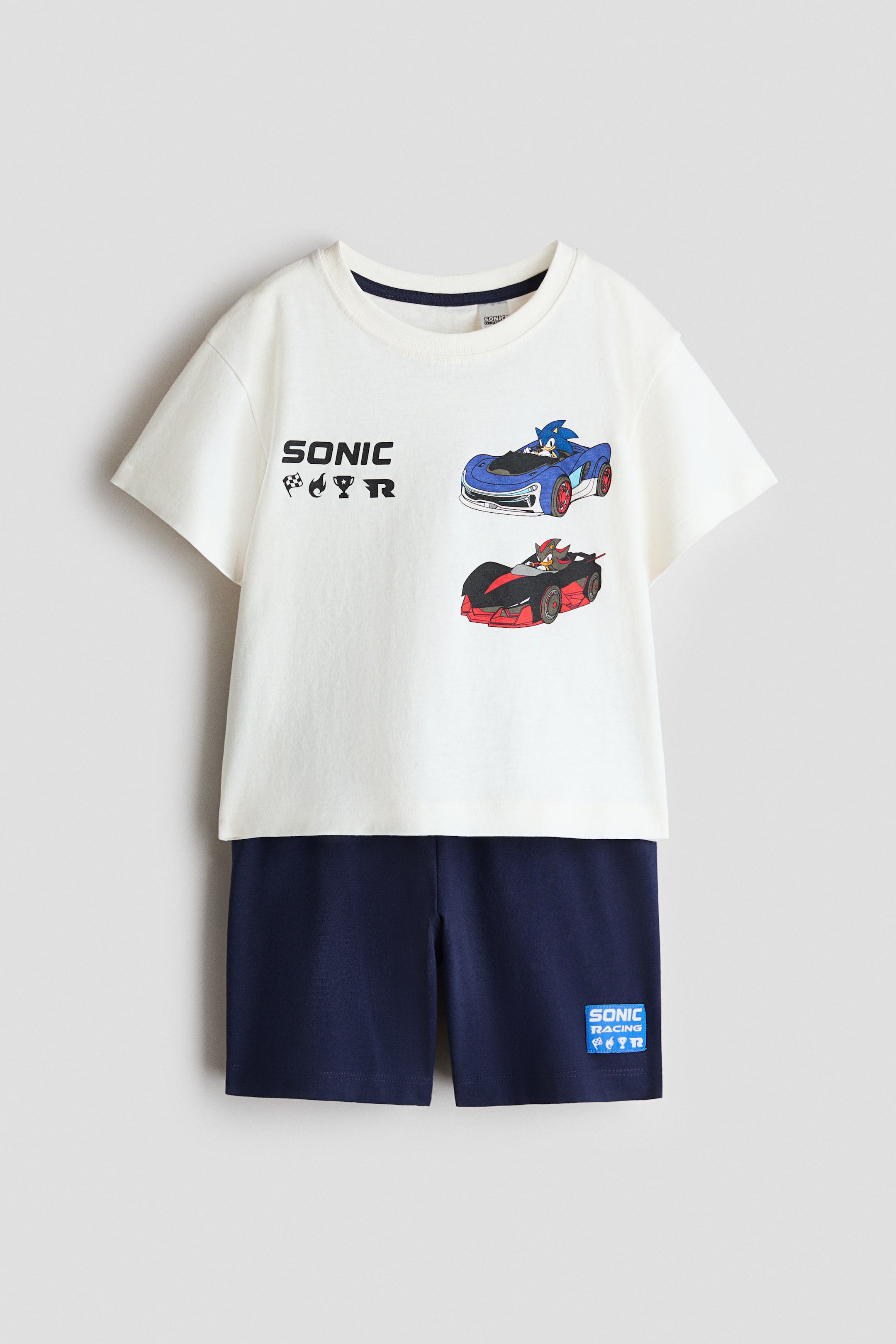 Vis større bilde: Pyjamas med en kremfarget T-skjorte og matchende marineblå shorts ligger flatt, vendt mot fronten. T-skjorten har en ribbestrikket kant rundt halsen, lett droppede skuldre, og et fronttrykk som viser teksten 'SONIC' med racingikoner, samt Sonic og Shadow i racerbiler, mens de marineblå shortsene er designet med en elastisk linning og en 'SONIC RACING'-patch på høyre ben.