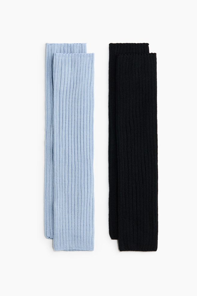Dos pares de calentadores de piernas de tejido acanalado estÃ¡n colocados extendidos uno junto al otro, mostrando su textura vertical acanalada y su diseÃ±o recto y tubular. El par de la izquierda es azul claro y el par de la derecha es negro; ambos estÃ¡n orientados al frente para mostrar su estructura. - 1