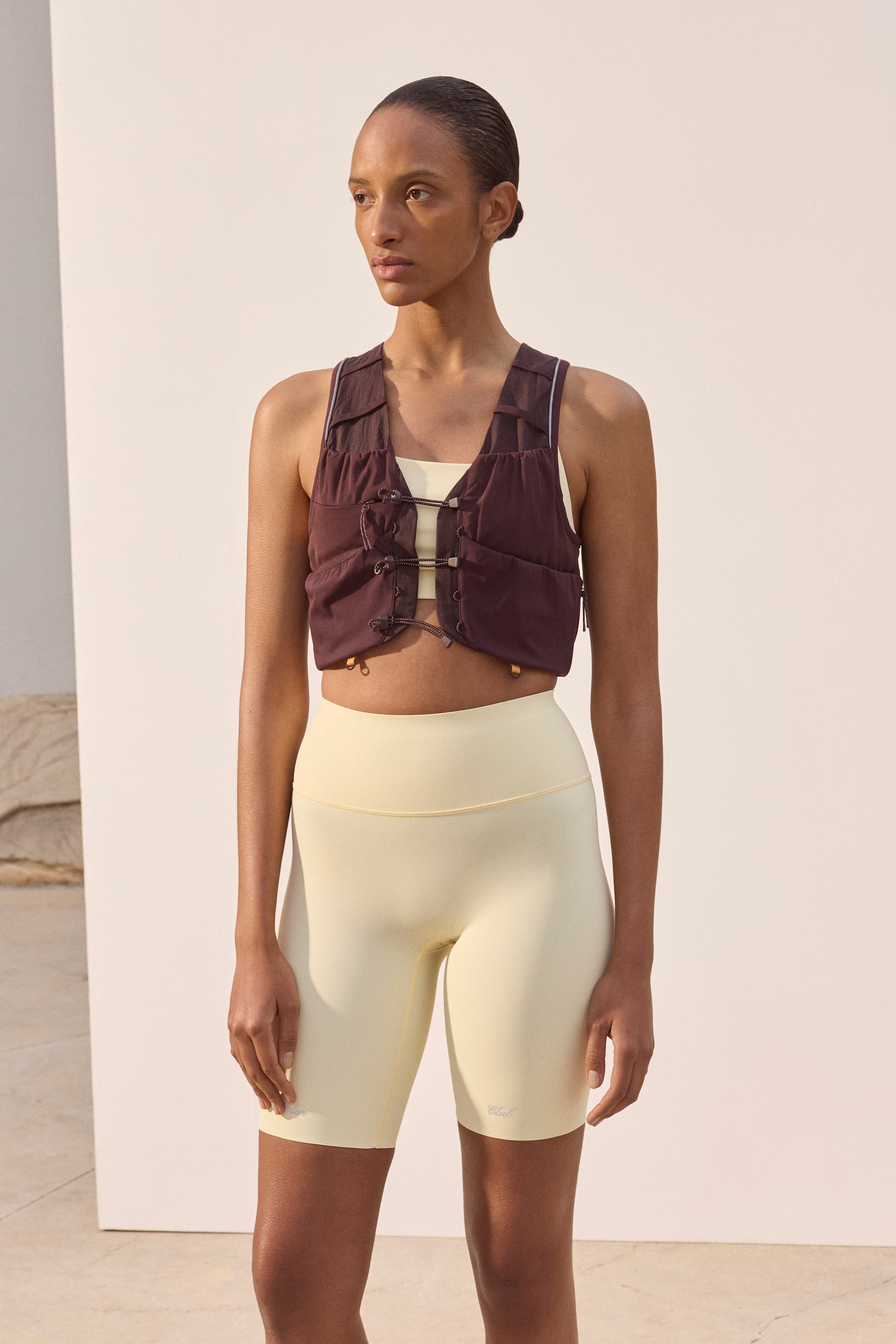 View larger image: A dark brown cropped utility vest with ruched details, drawstrings, and toggles is being layered over a light yellow sports bra, and is being paired with high-waisted, pale yellow biker shorts. The vest features thin white piping along its shoulder straps, and the biker shorts display a subtle logo on the left thigh.